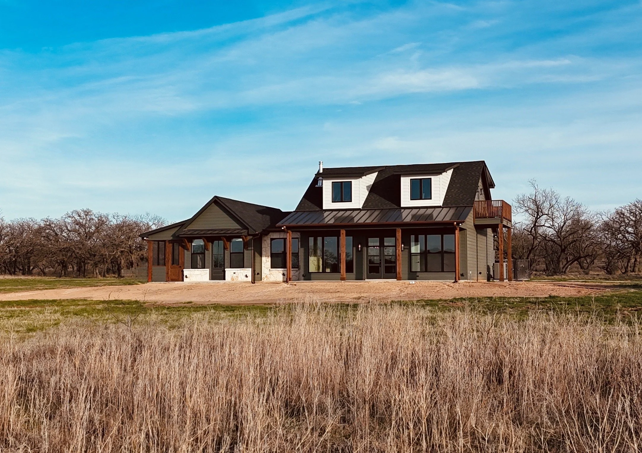 A modern two-story house with large windows, dark roof, and wooden accents, situated in a rural area with dry grass and leafless trees in the background under a clear blue sky, built by Lowrance Signature Homes.