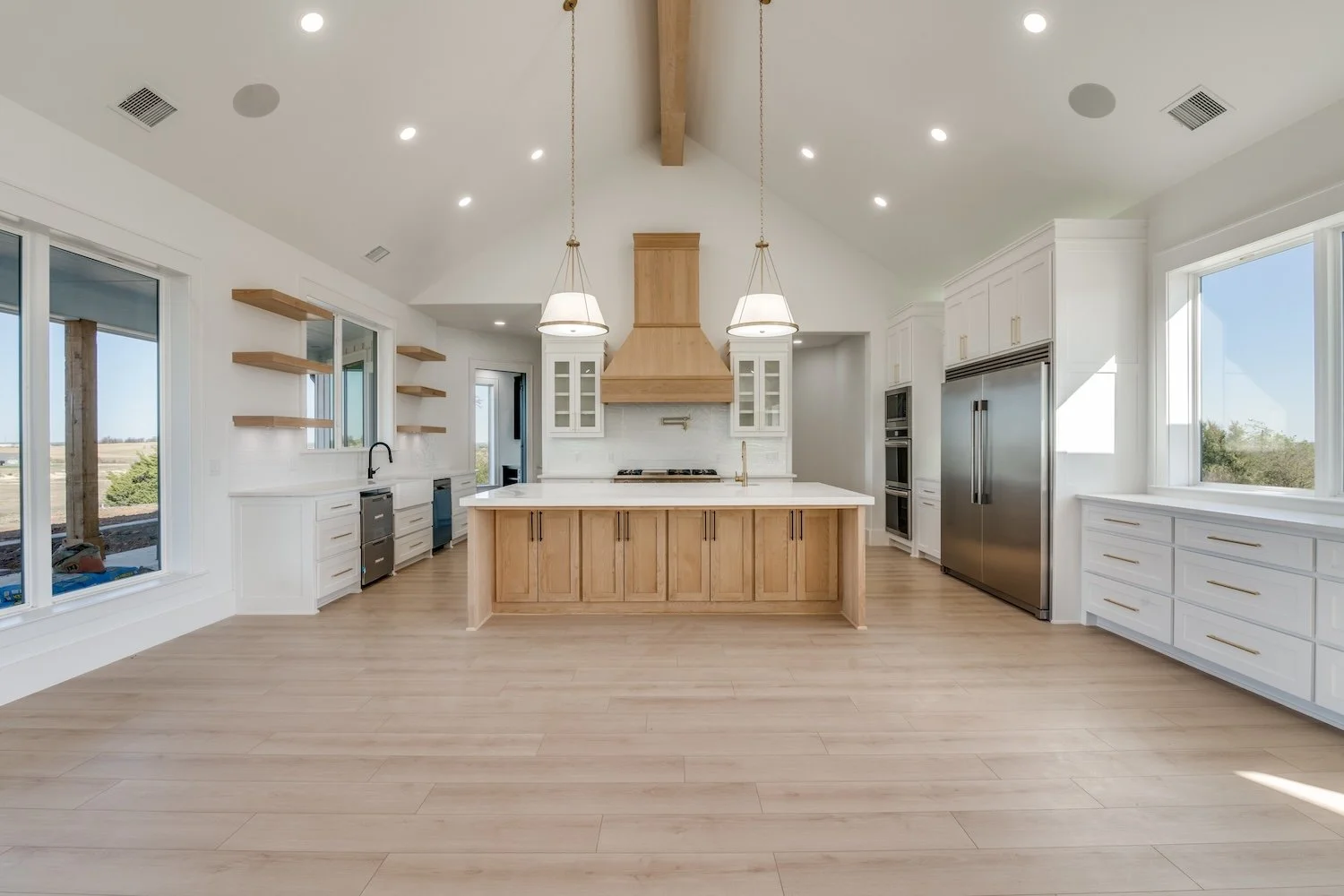 Modern white and light wood kitchen with large central island, stainless steel refrigerator, and open shelves. Multiple windows let in natural light, and there are ceiling lights and two pendant lights over the island.