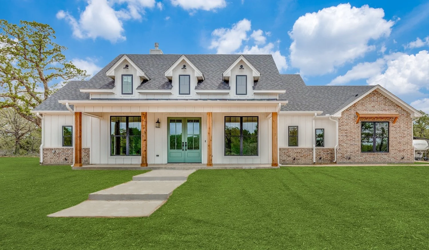 Front view of a modern house with a gray shingled roof, white vertical siding, brick accents, and turquoise double front doors. The house has three dormer windows and black-framed windows on either side of the doors, with a concrete pathway leading to the entrance. The lawn is green and well-kept, with a large tree in the background under a partly cloudy sky.
