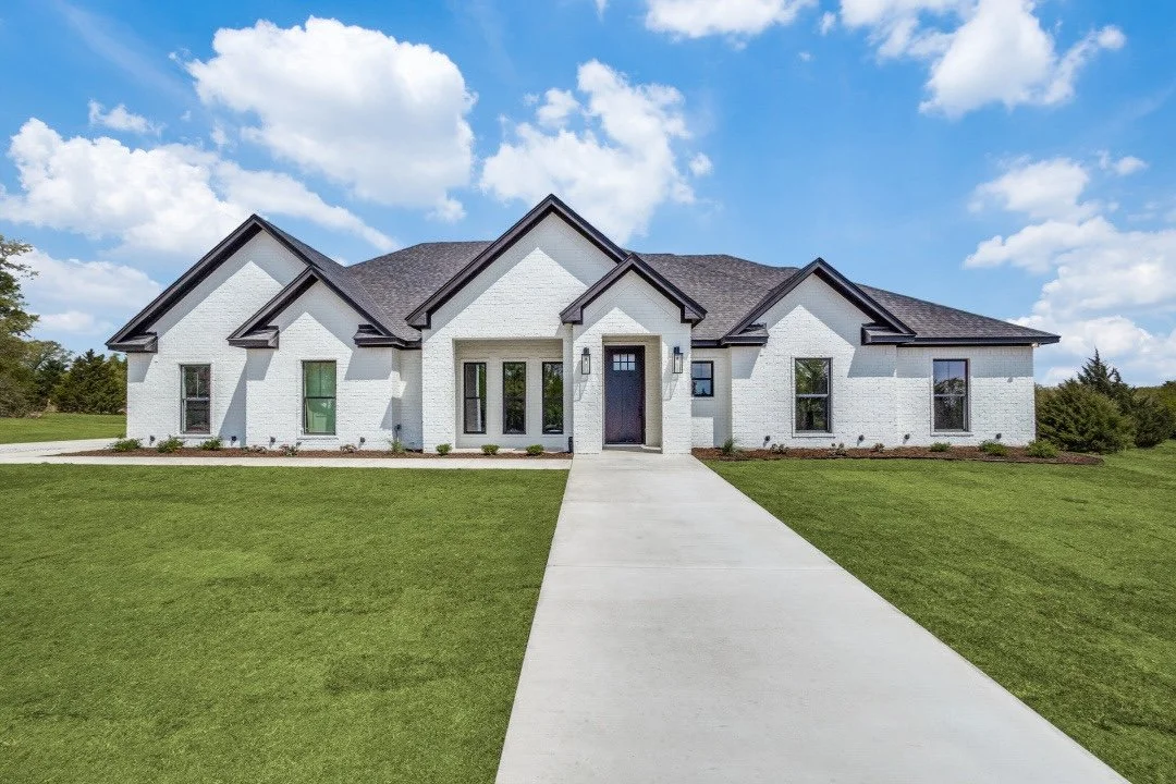 Front view of a large white house with dark roof and multiple gables, concrete walkway leading to the front door, on a well-manicured lawn with a partly cloudy sky in the background.