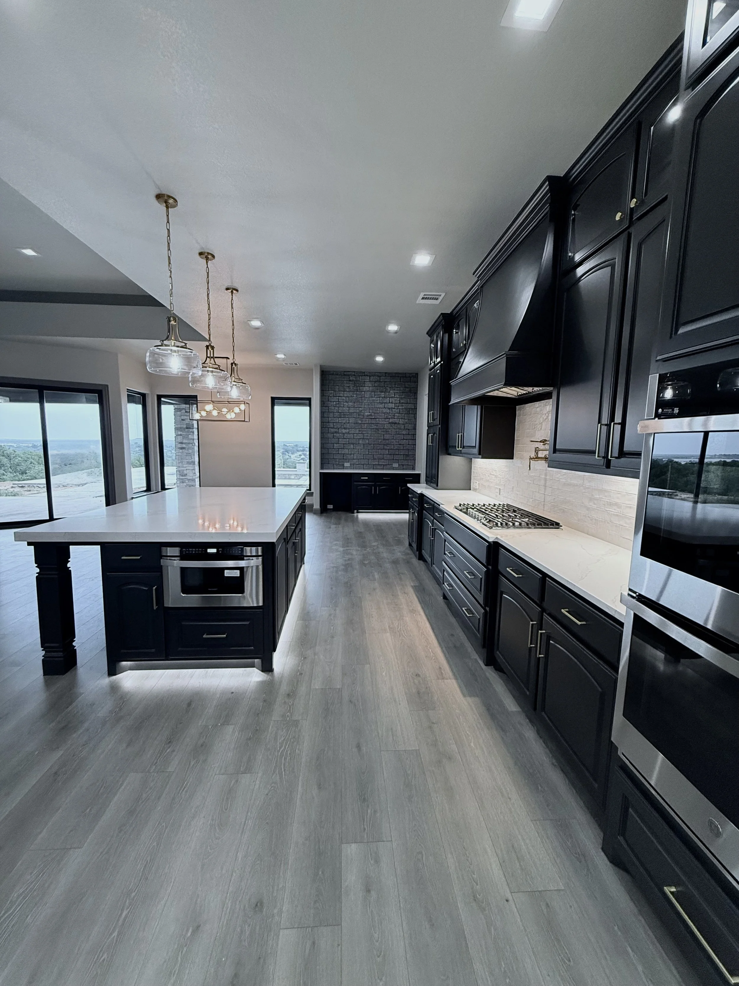 Modern kitchen with black cabinets, white countertops, gray wood flooring, and large windows showing a view outside, built by Lowrance Signature Homes.