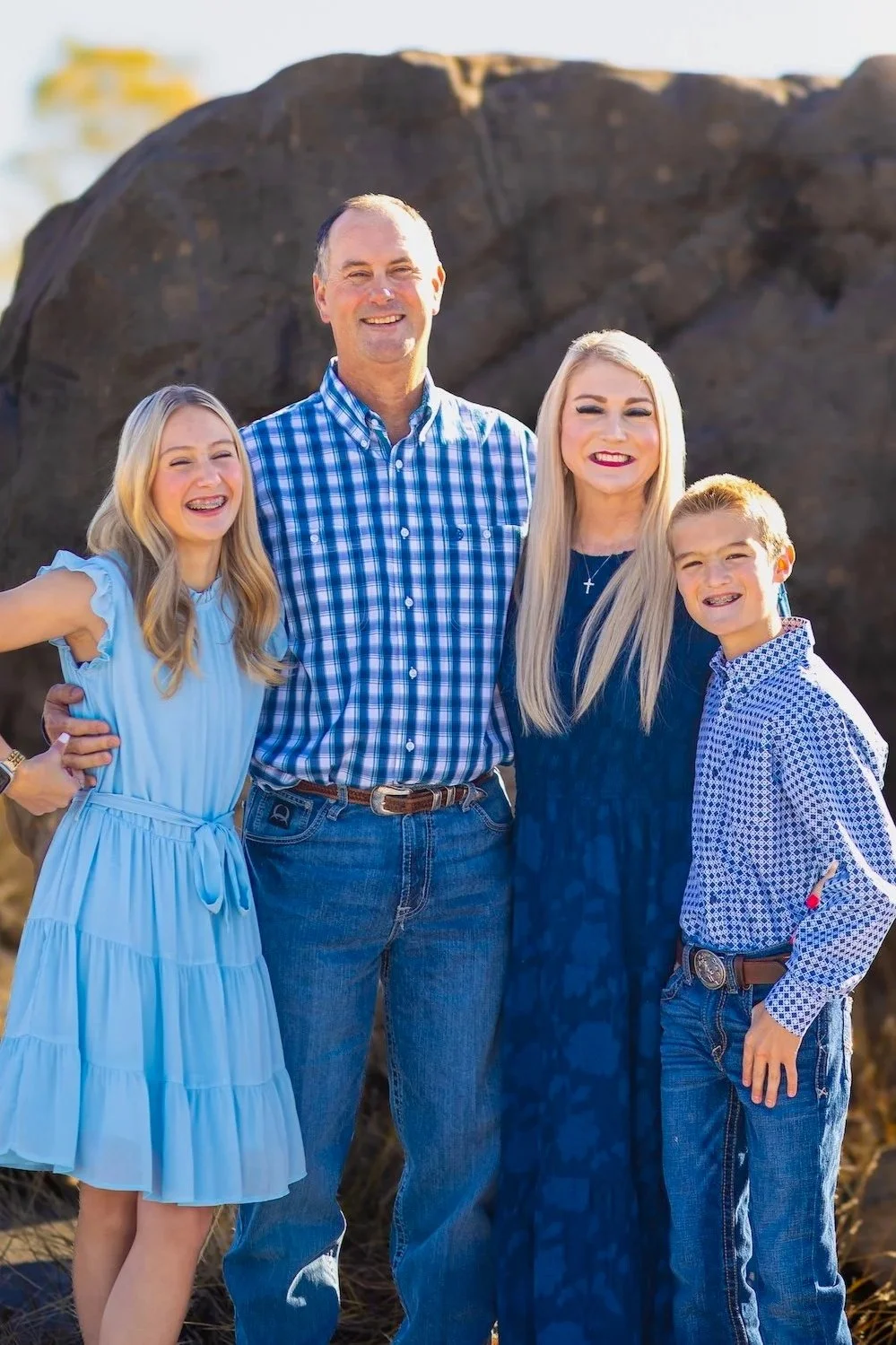 A family of four standing outdoors in front of a large rock, smiling at the camera. The man and woman are in the center, with the girl on the left and the boy on the right. They are dressed in casual, coordinated blue clothing.