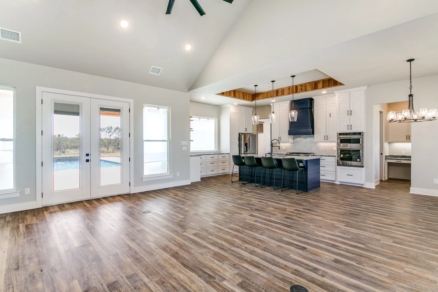 Open-concept kitchen and living area with hardwood floors, white cabinets, black island, hanging pendant lights, and large windows with a view of a pool outside, built by Lowrance Signature Homes.