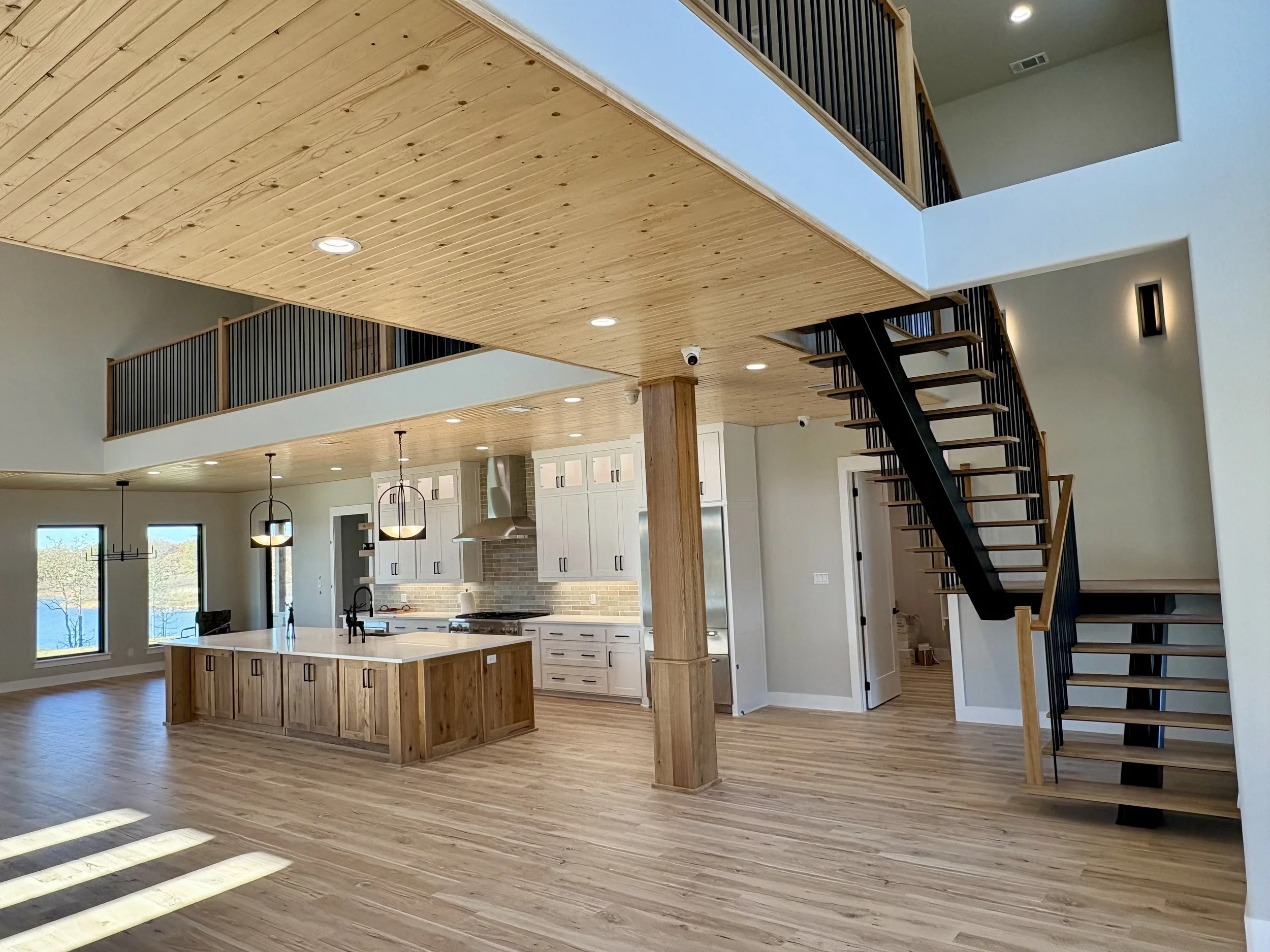 Open-concept kitchen and living area with wooden floors, a kitchen island, modern pendant lights, white cabinets, and a staircase leading to an upstairs loft, built by Lowrance Signature Homes.