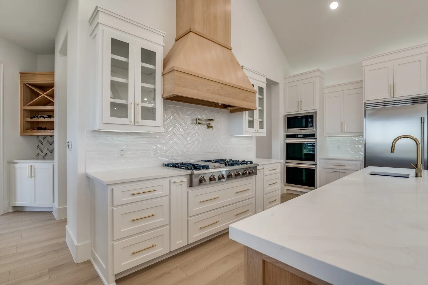 Modern kitchen with white cabinets, a gas stove with brass knobs, a large wooden range hood, and built-in microwave and oven. There is a white marble countertop island with a brass faucet, and a wine rack is visible on the left side.
