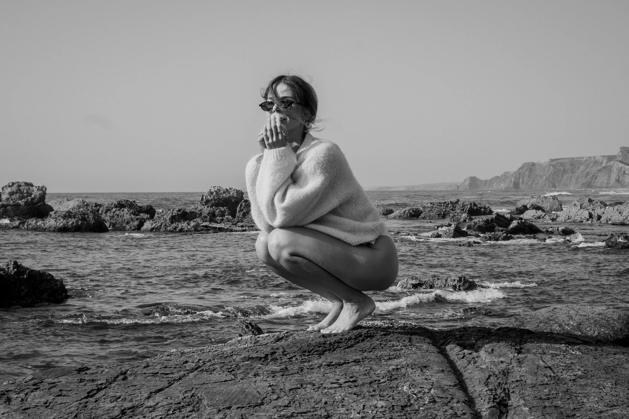 A woman wearing sunglasses and a chunky sweater crouching on rocks by the sea, with cliffs in the background, in black and white.