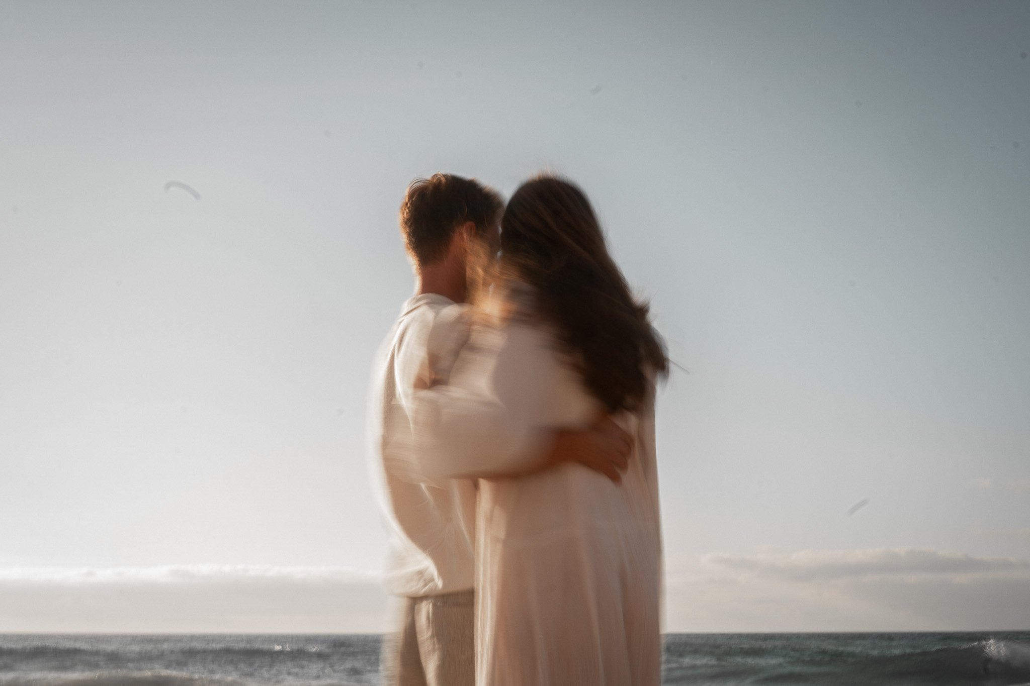 A couple standing on a beach facing each other at the shoreline, with the ocean and sky in the background.
