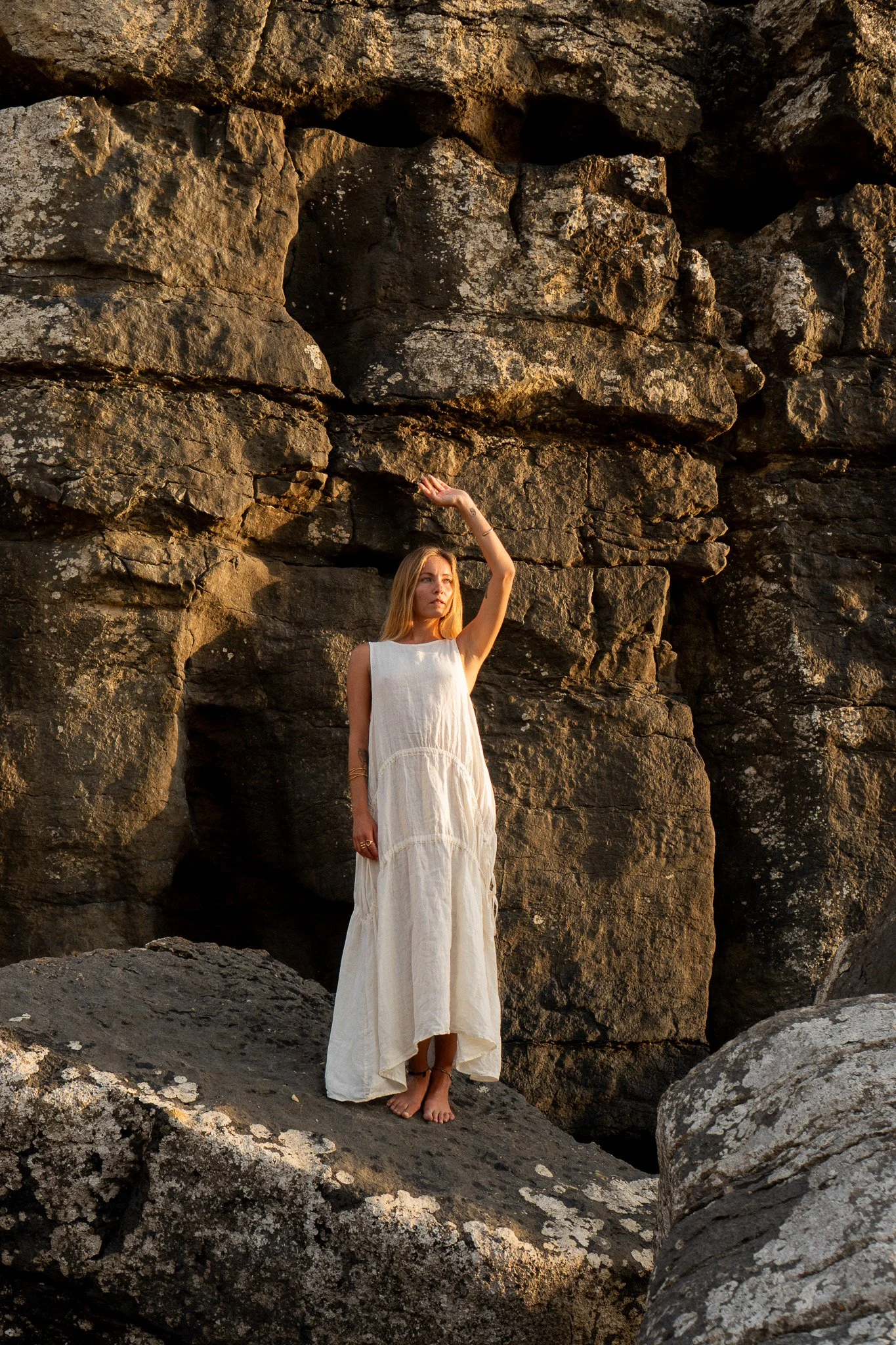 A woman in a white dress barefoot standing on rocks next to a large rocky formation, with her right arm raised and hand touching the rocks, during sunset or golden hour.