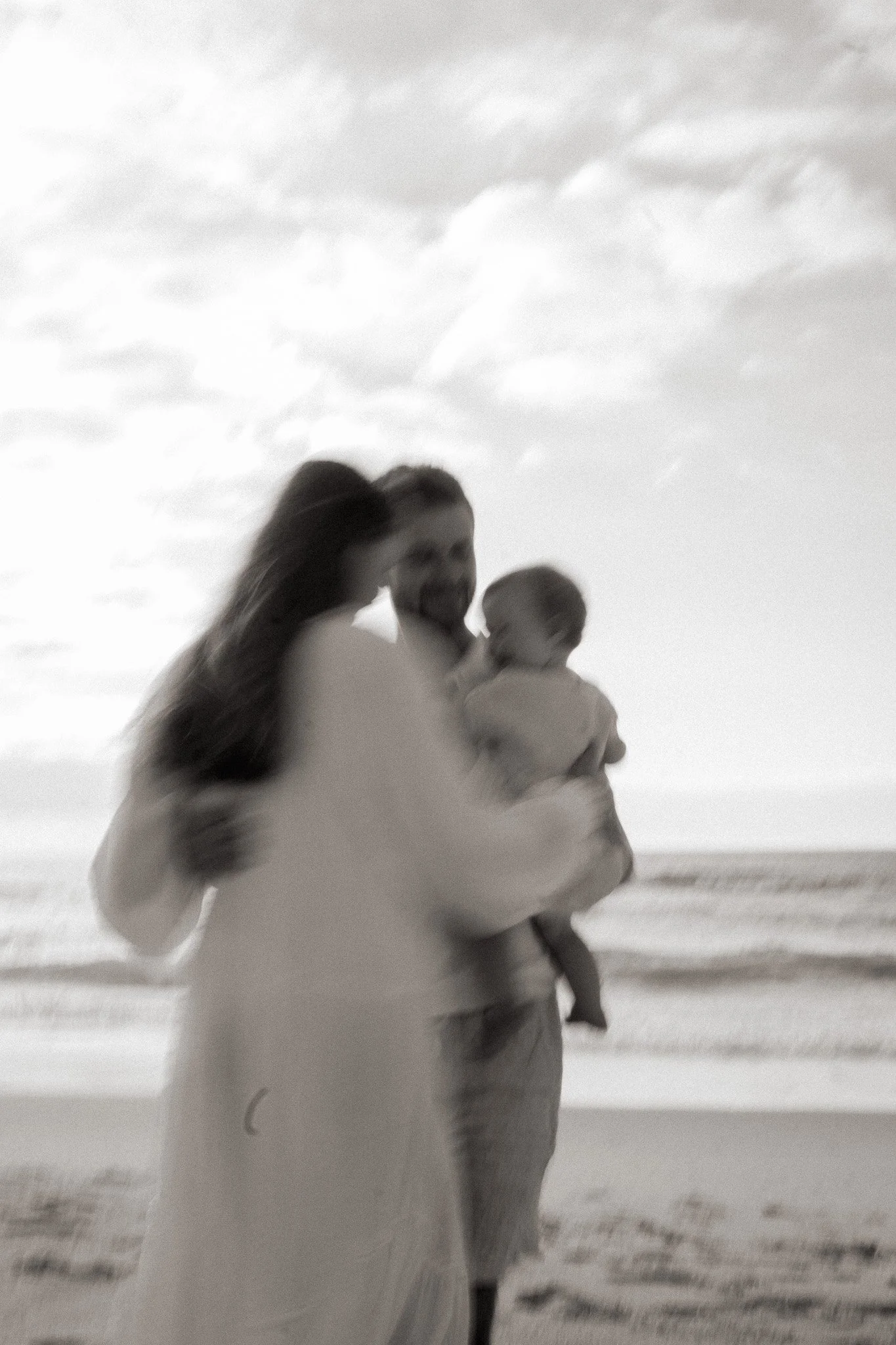 A woman holding a child on a beach, with another person in the background, under cloudy sky.