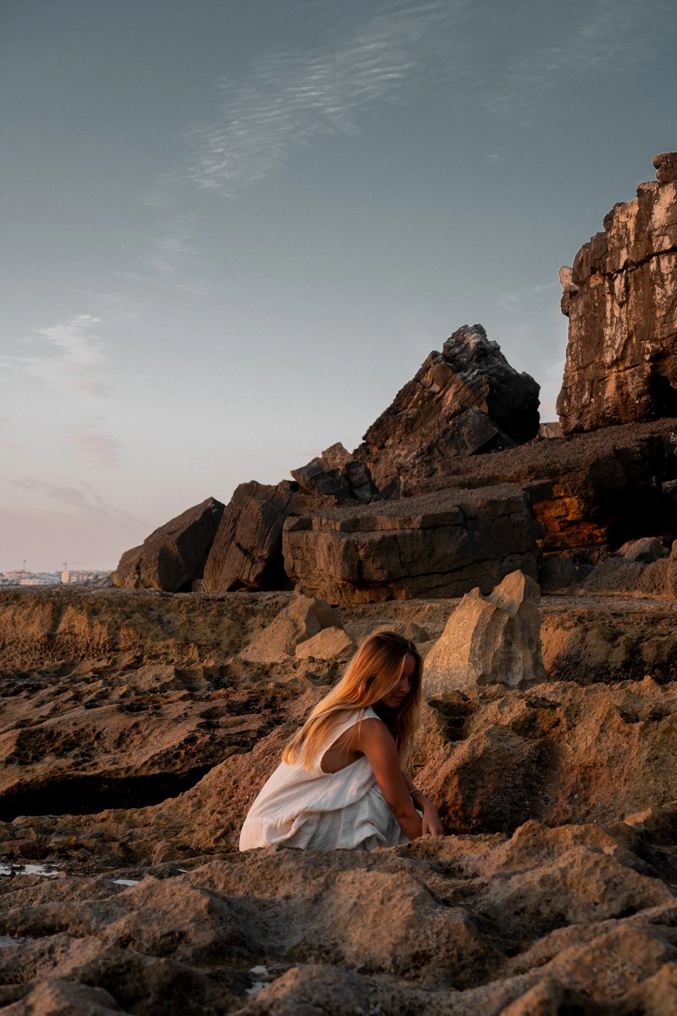 A woman with long blond hair in a white dress sitting on a rocky terrain near large rock formations at sunset or sunrise.