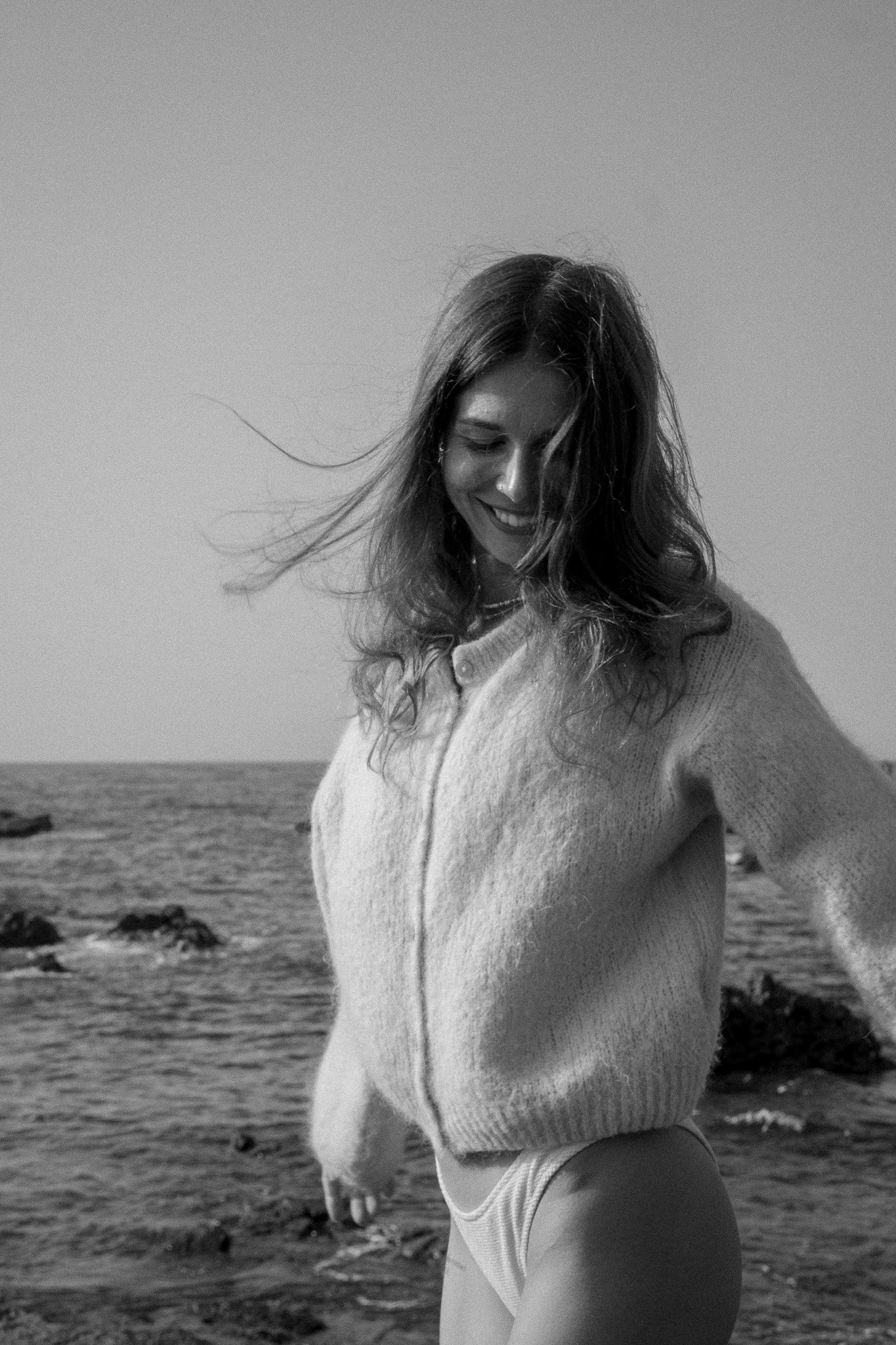 A woman smiling at the beach, looking down, with wind-blown hair, wearing a sweater and shorts, standing near the ocean with rocks in the background.