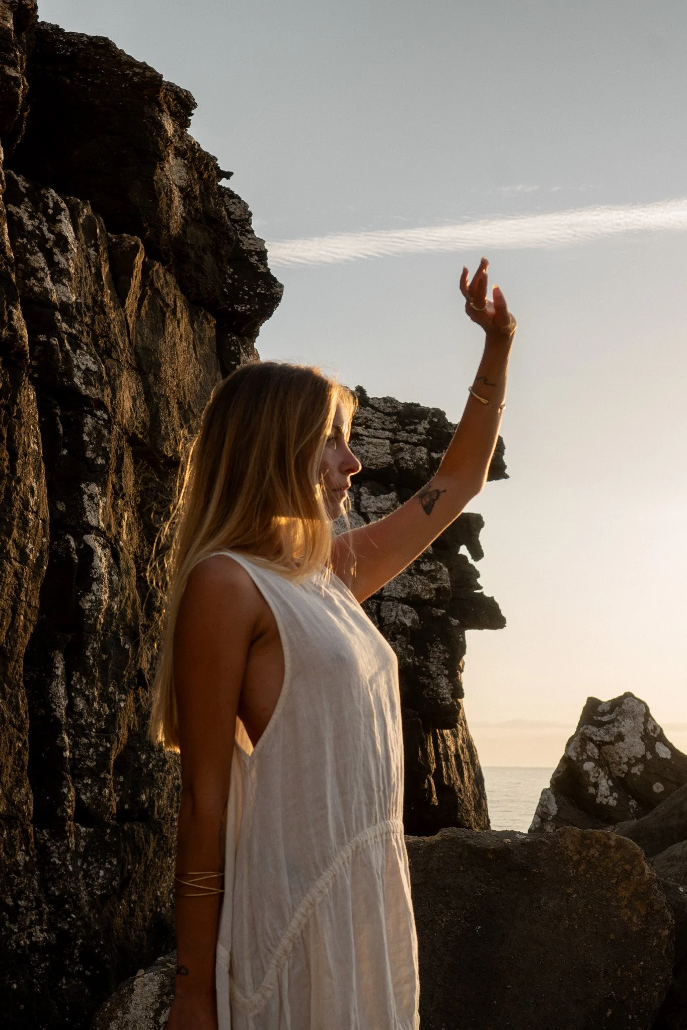 A woman in a white dress standing near rocky cliffs by the sea during sunset, with her arm raised.