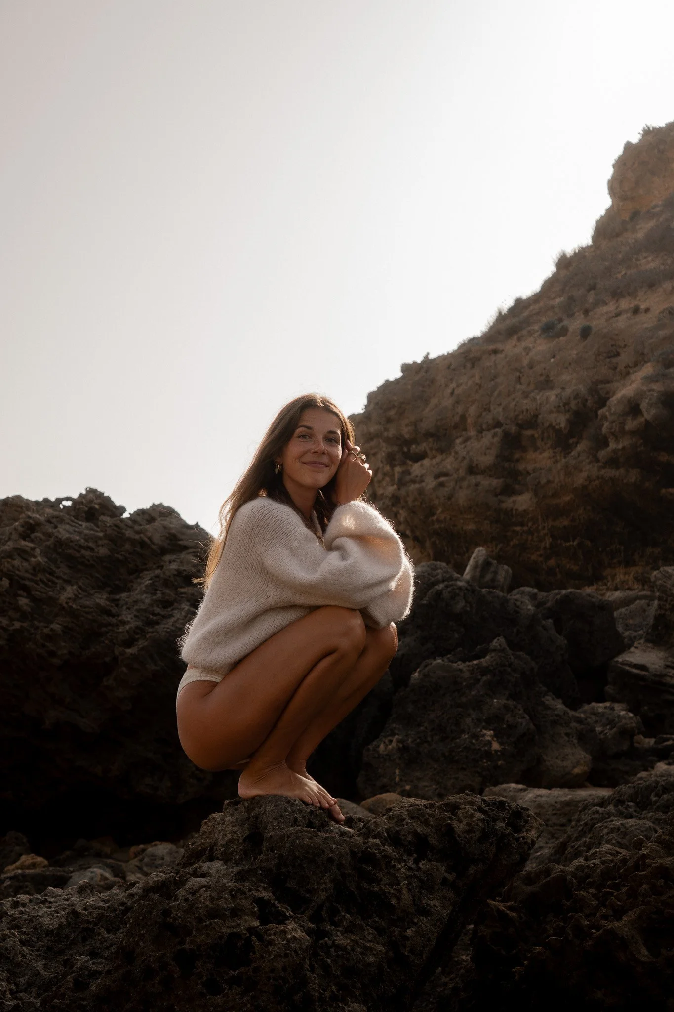 A woman with long brown hair sitting on rocks, smiling, with a rocky hillside and sky in the background.