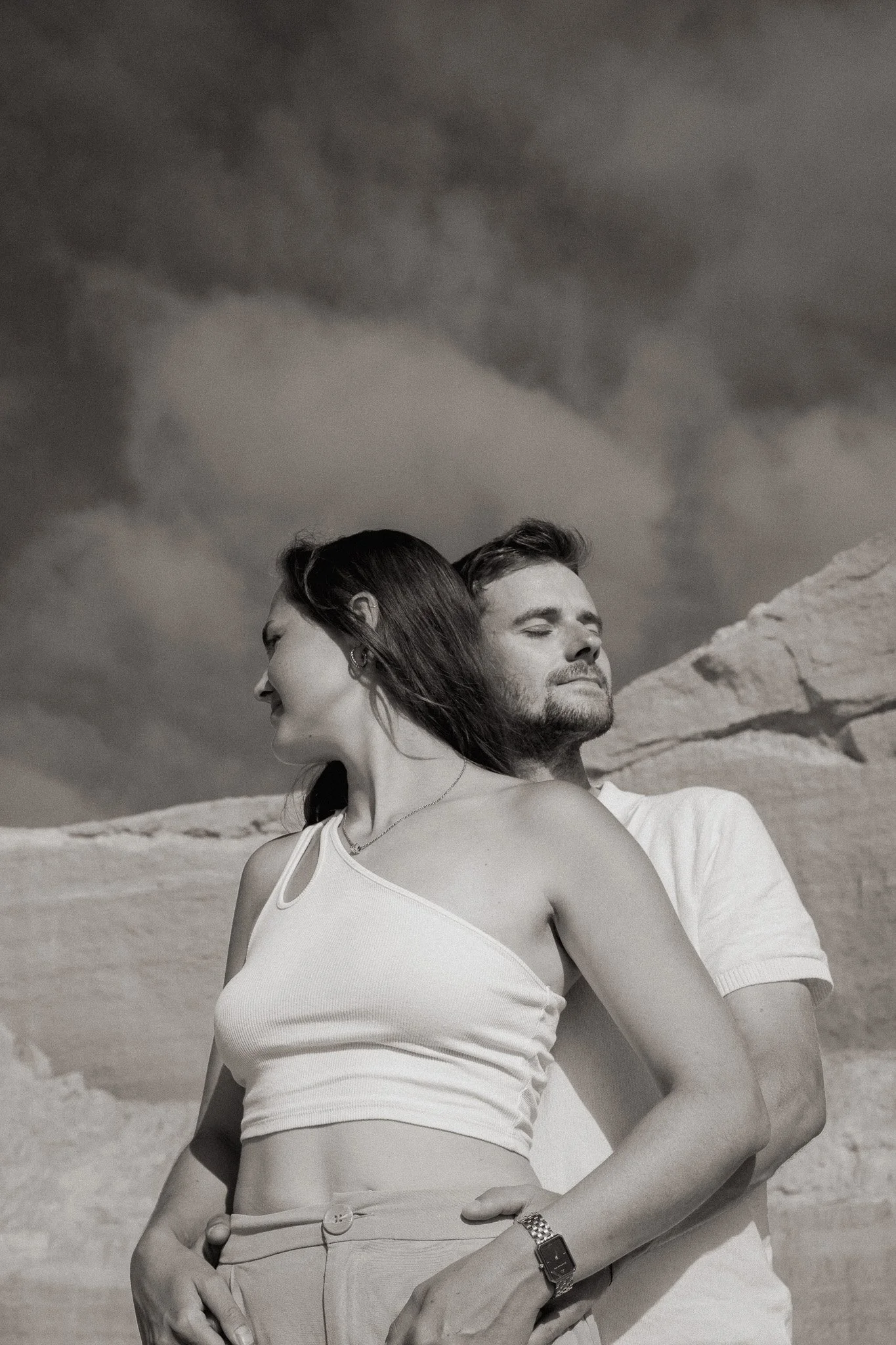 A man and a woman standing outdoors in front of a cloudy sky. The woman is in the foreground, wearing a sleeveless top and high-waisted pants, looking to her left. The man, standing behind her, has his eyes closed and appears to be relaxed. The background includes a rocky terrain.