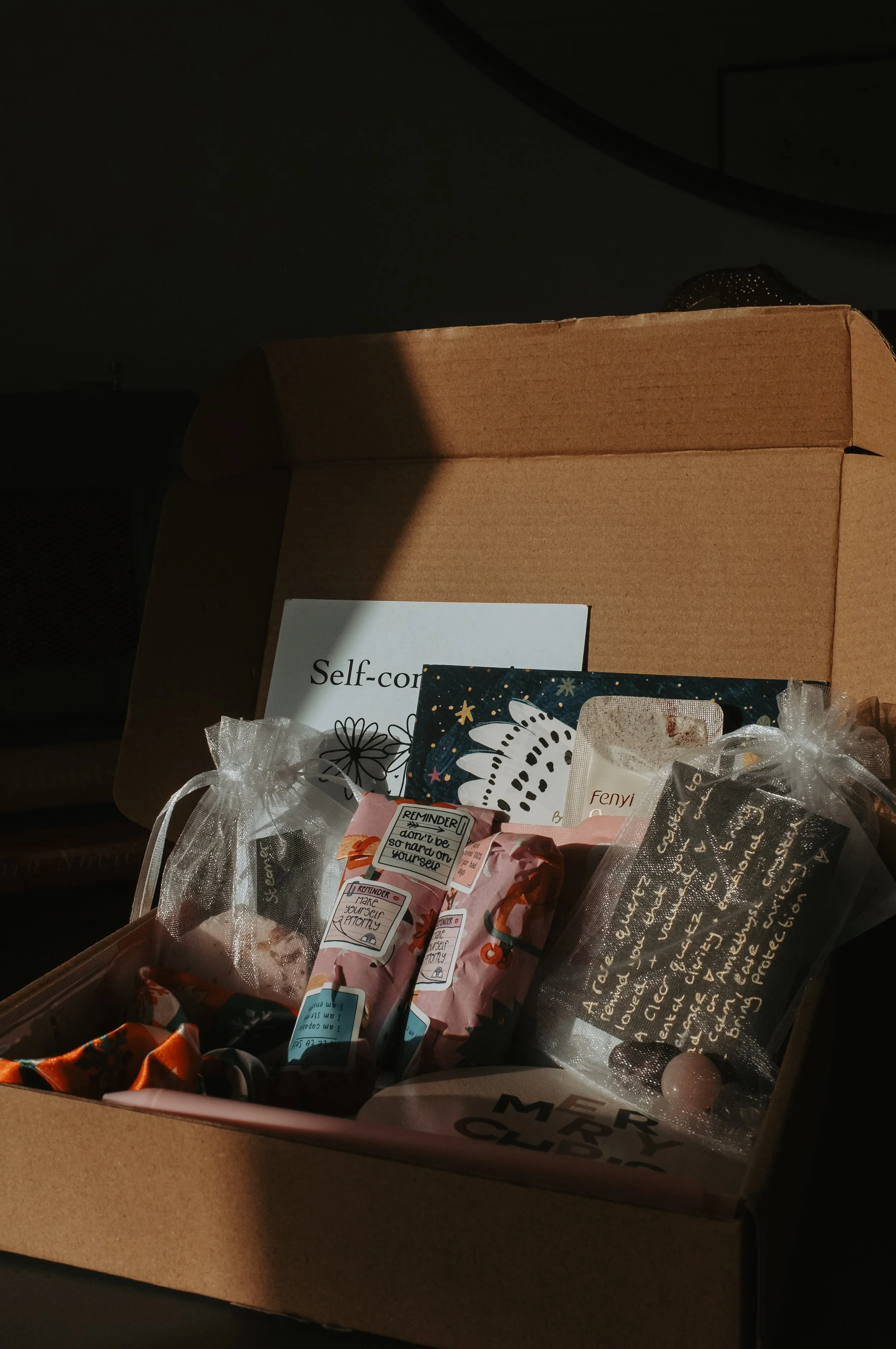 Open cardboard box filled with various gift items, including small bags, cards, and a note, with a partial shadow cast over part of the box.