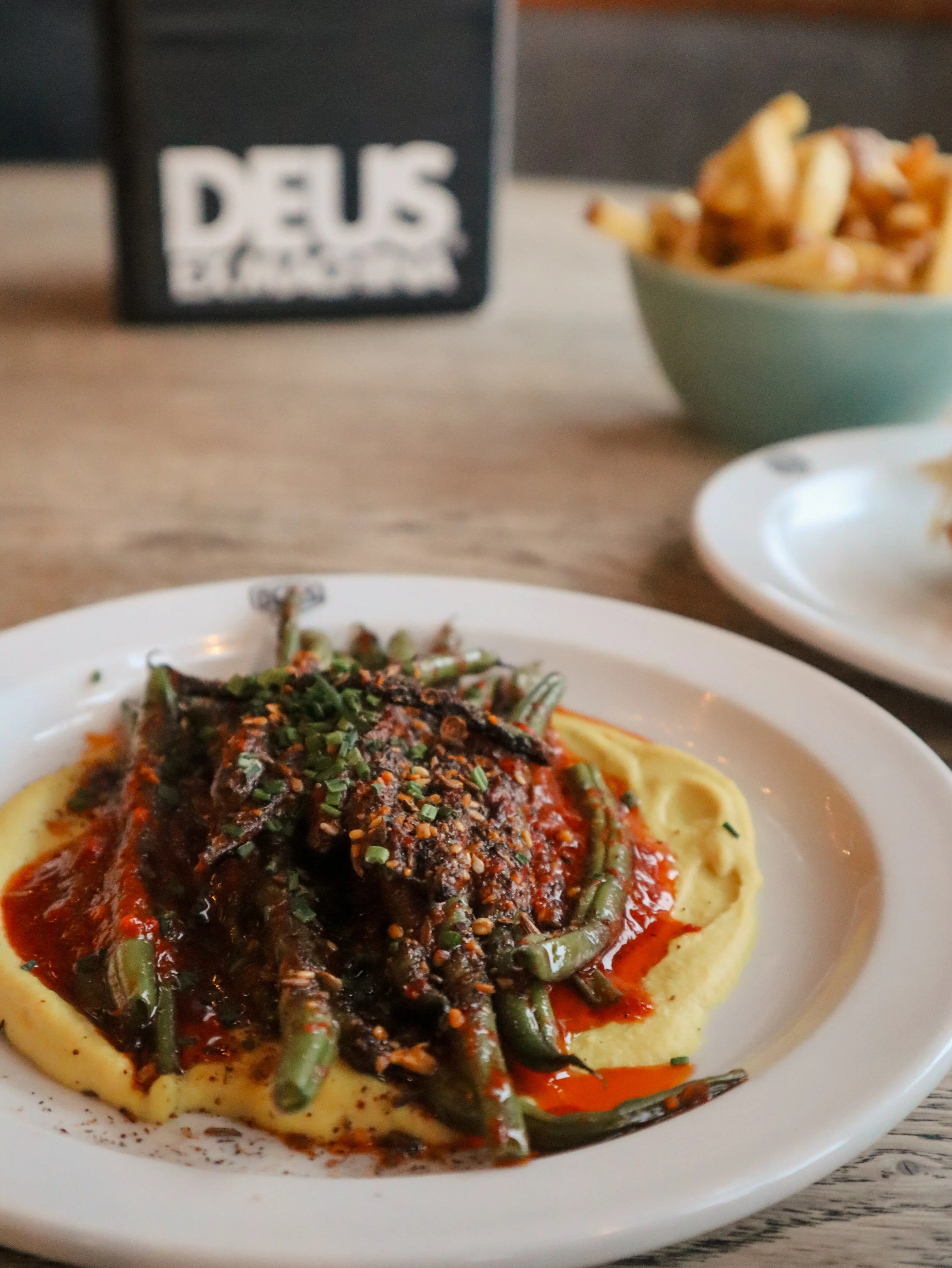 A plate of pasta topped with a red sauce and herbs, with green beans as a side, on a white plate with a panda design, on a wooden table. In the background, there is a bowl of French fries and a blurry sign that says 'DEUS.'