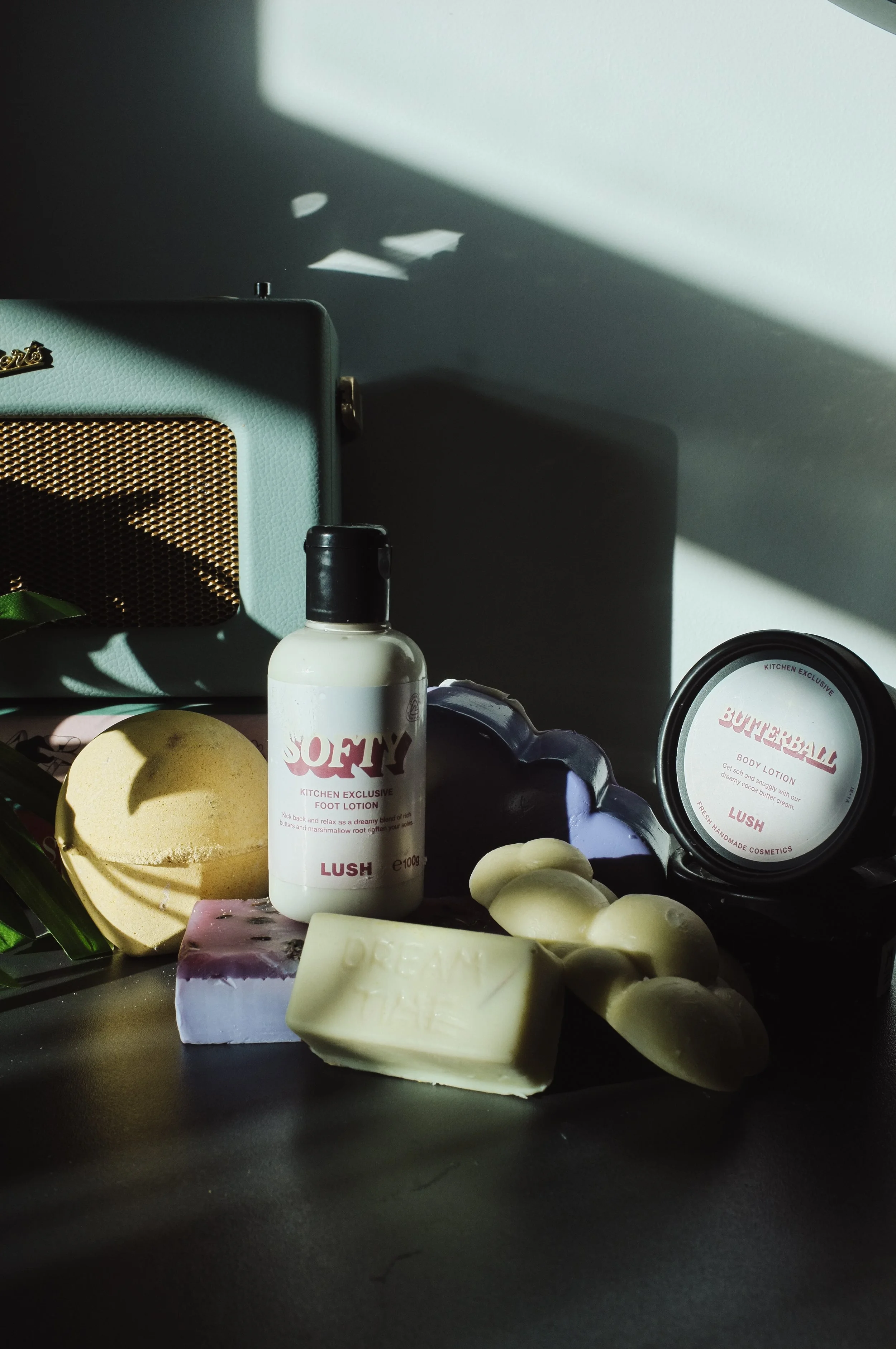Lush skincare products, including foot lotion, body lotion, soap, and bath bombs, arranged on a table with shadows and sunlight streaming in.