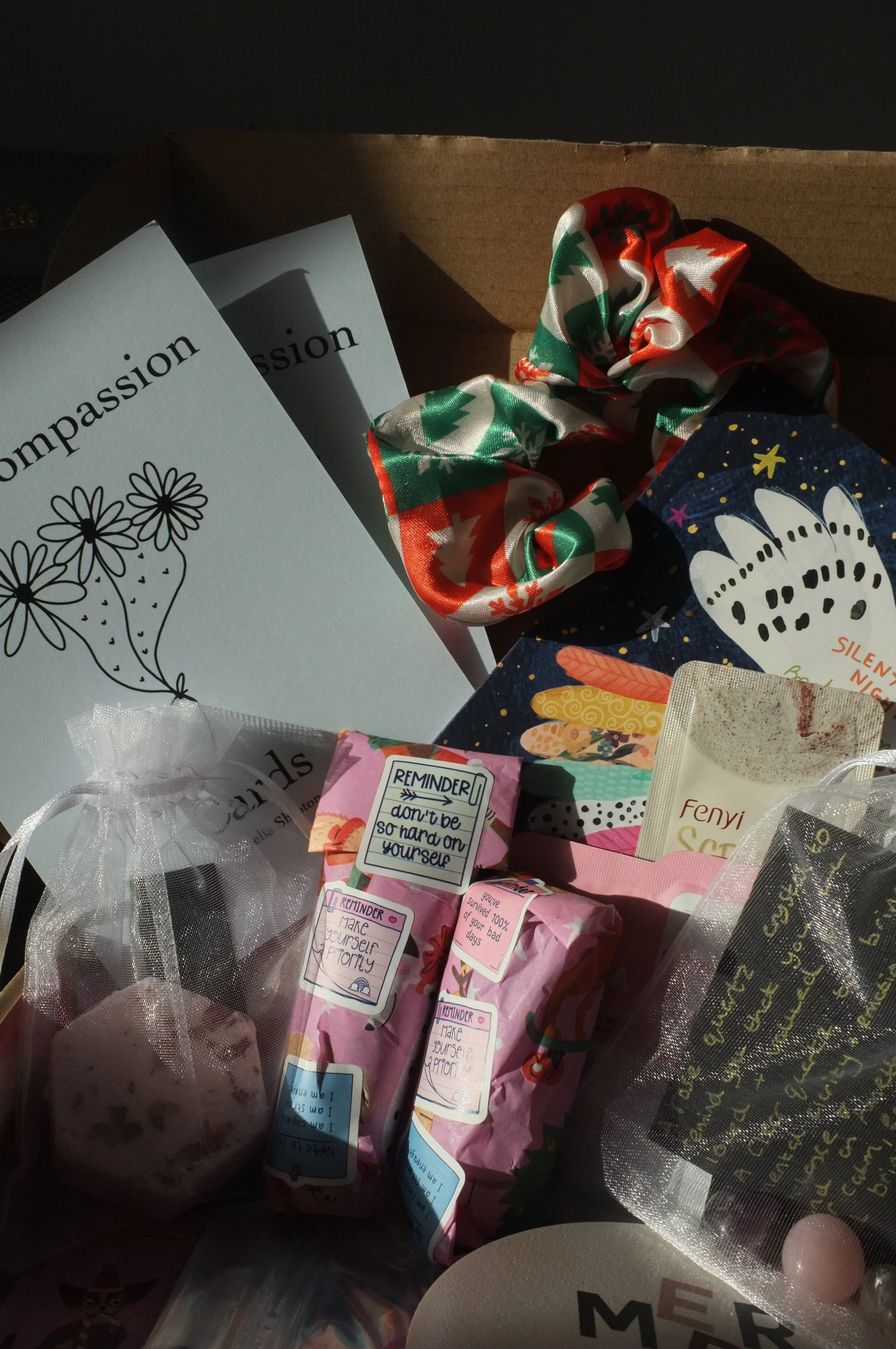 Holiday gift box containing a card with the word 'Compassion', a scrunchie with a Christmas pattern, wrapped soap, and various small gifts and notes.