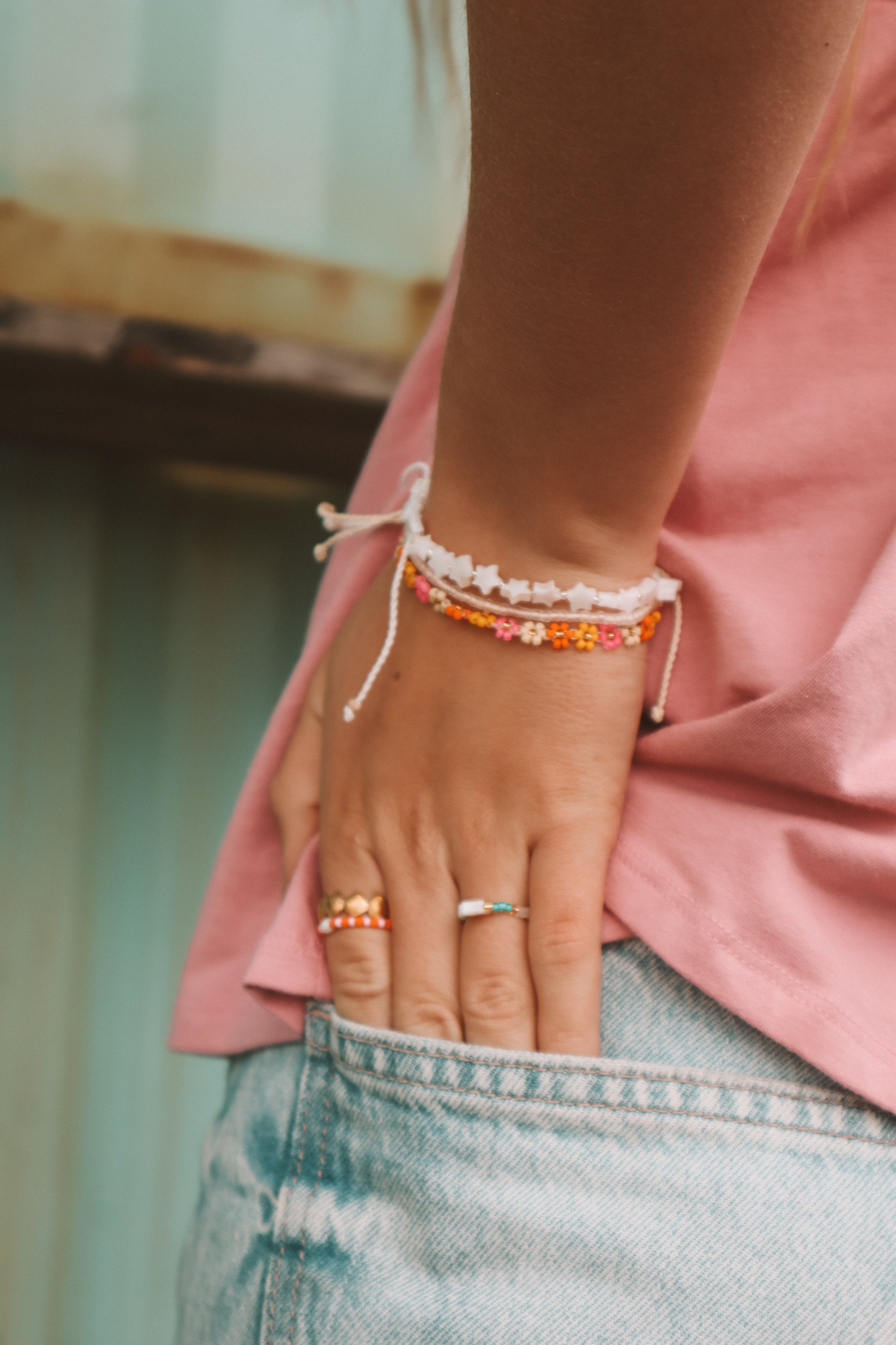 Close-up of a person's arm wearing colorful beaded bracelets and rings, with pink clothing and light blue denim shorts.