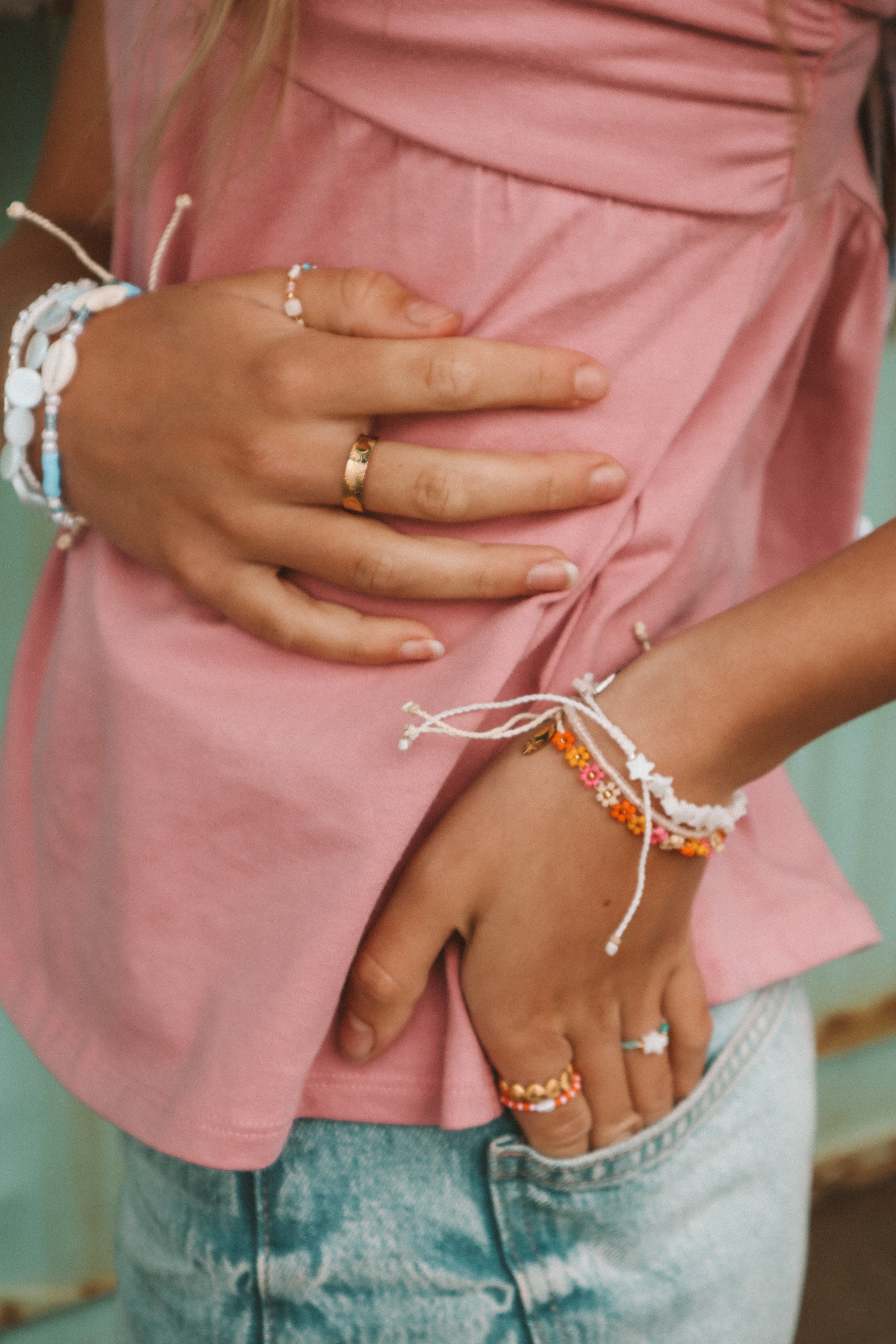 A person wearing a pink shirt and light blue jeans has their arms crossed over their chest. They are wearing multiple bracelets and rings on their fingers.