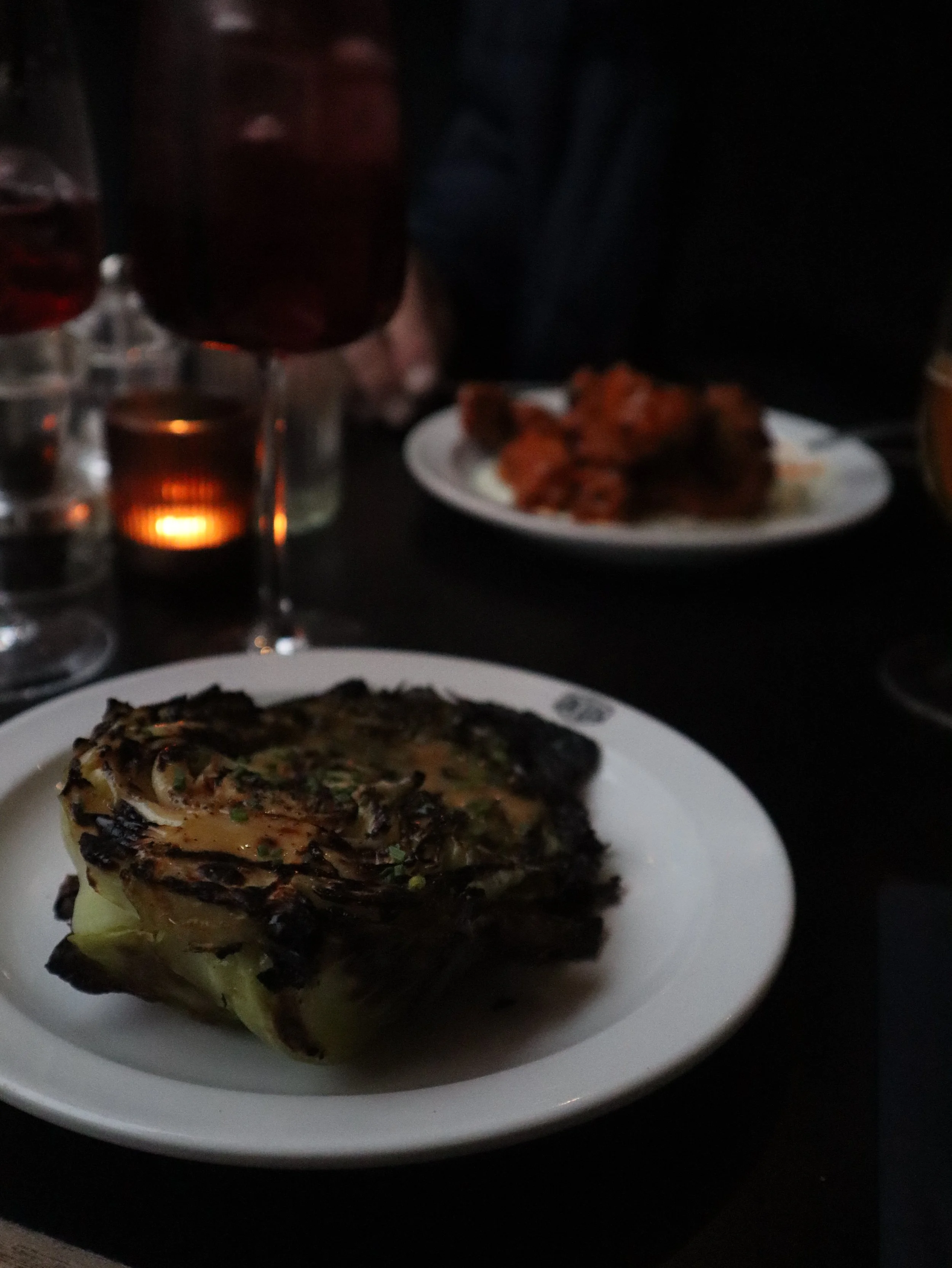 A dinner table with a grilled fish fillet on a white plate in the foreground, a candle holder with a lit candle, glasses of beer, a glass of red wine, a plate of fried chicken wings, and a dark background.