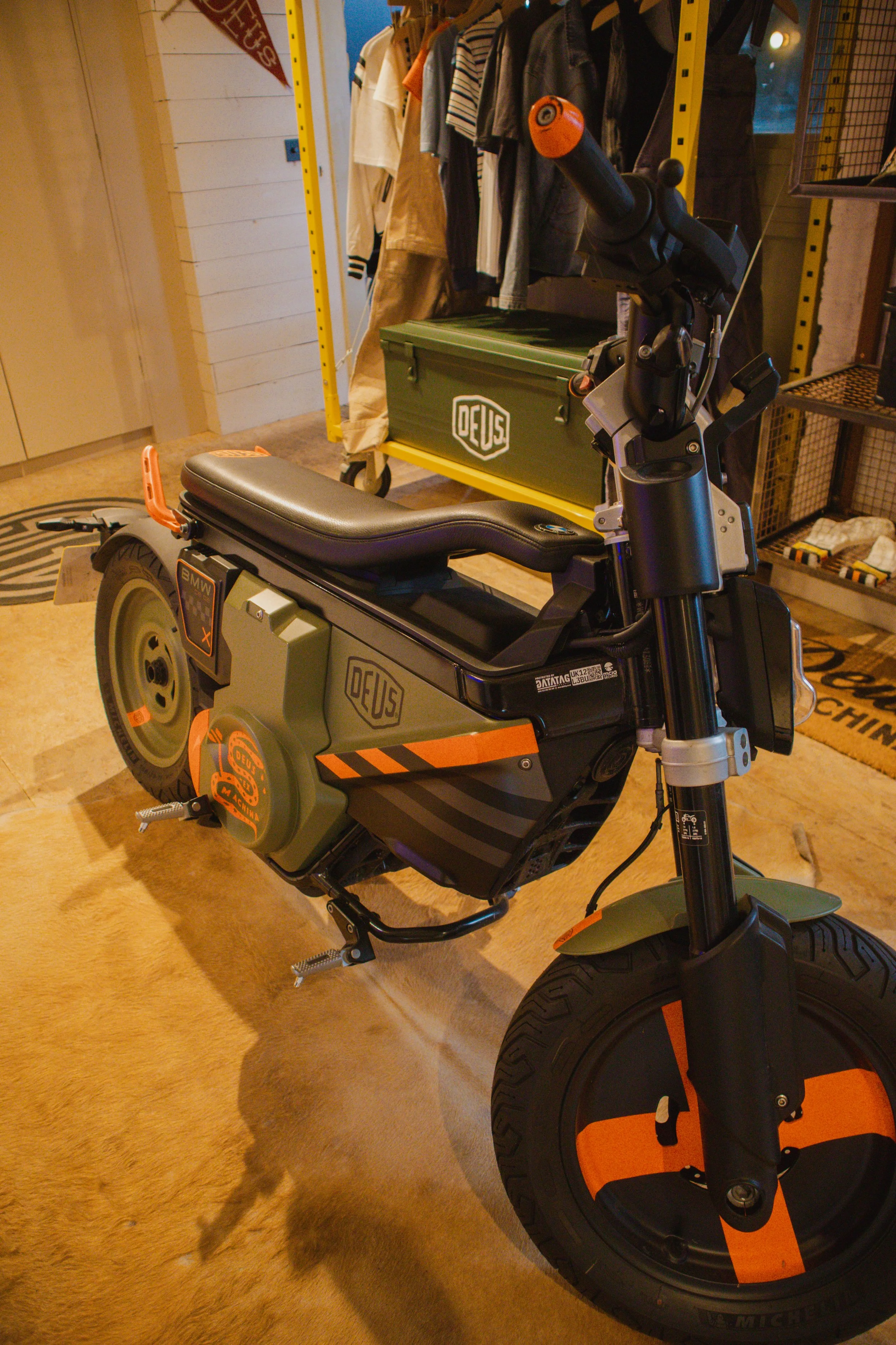 A small black and orange electric scooter parked indoors with a clothing rack, a green storage box labeled 'DEUS,' and a beige carpeted floor in the background.