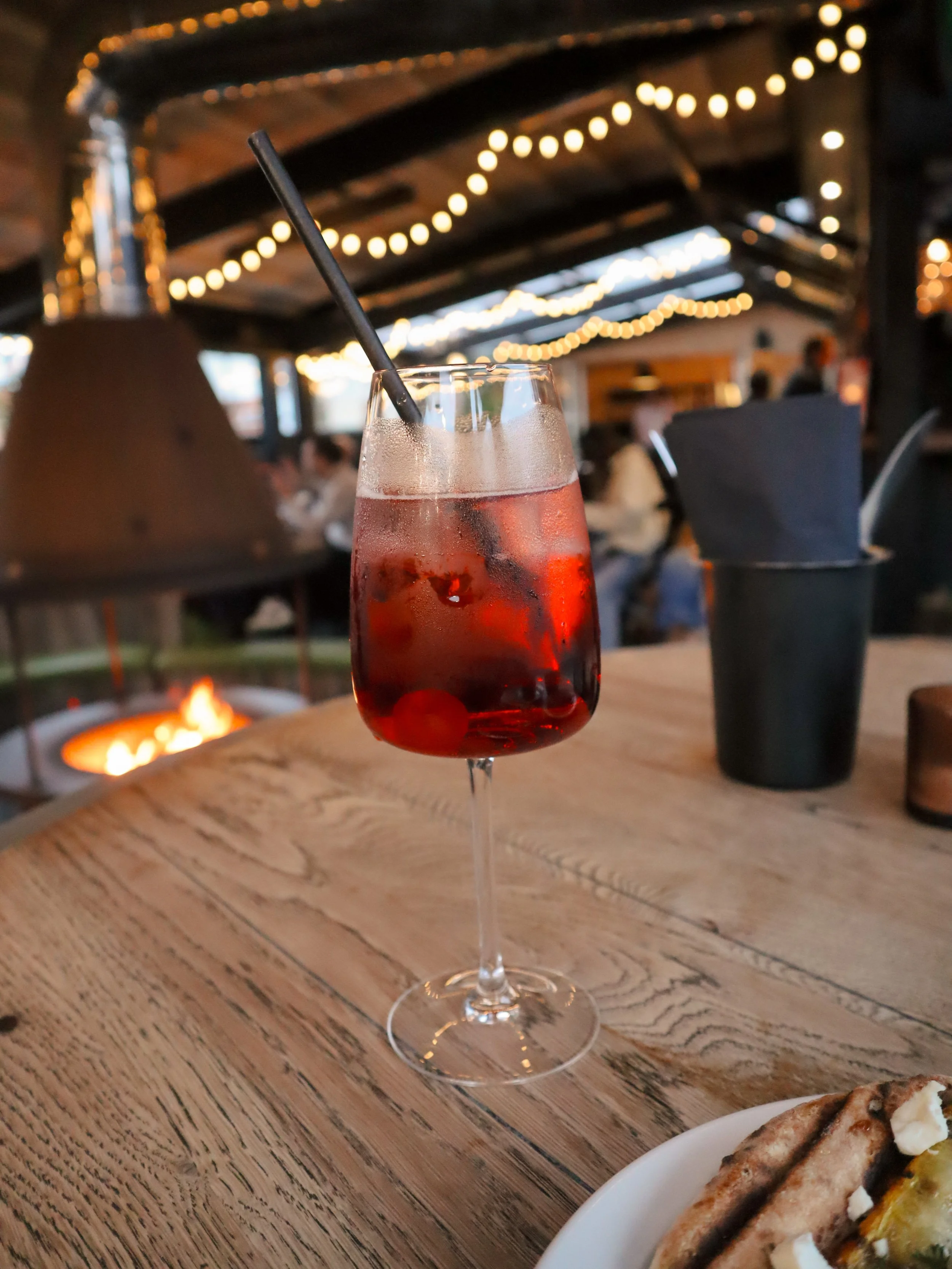 A glass of pink beverage with ice and a black straw on a wooden table, blurred background of an indoor dining area with string lights and people.