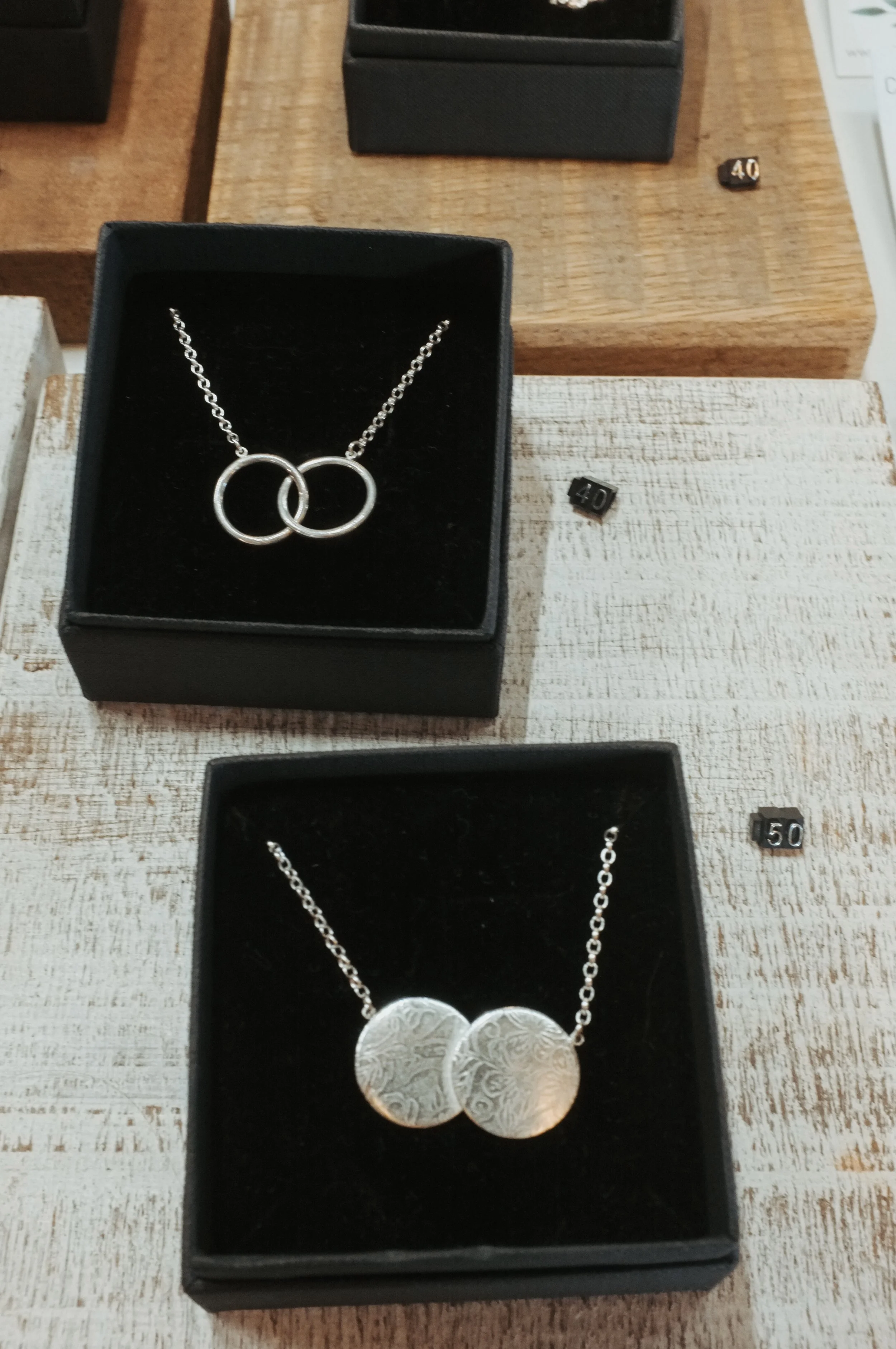 Two necklaces in black jewelry boxes displayed on a wooden table. The top necklace has two interlinked rings, and the bottom necklace has two textured circular pendants.