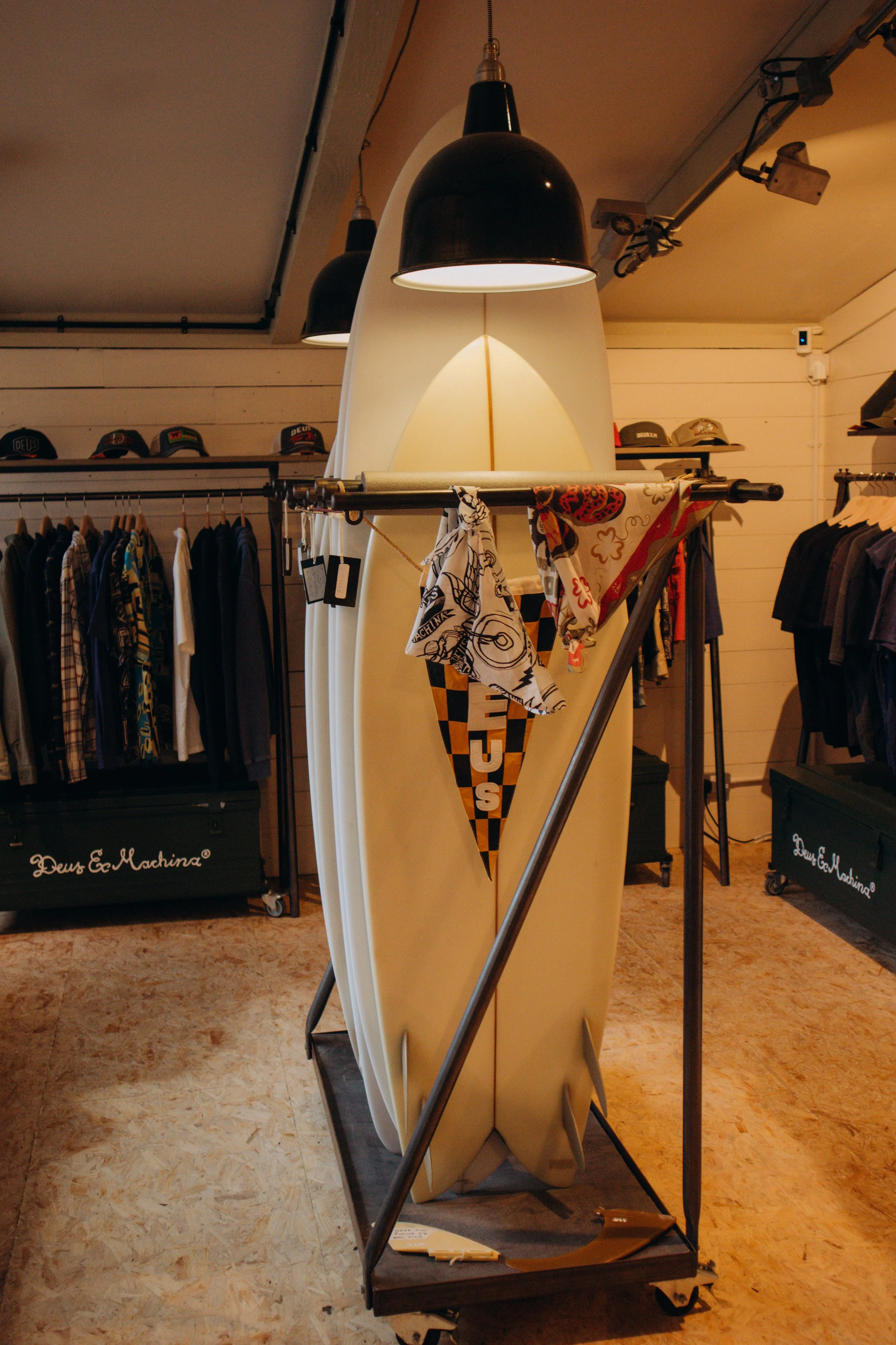A yellow surfboard resting upright on a wheeled display stand in a clothing store, with clothing racks and hats visible in the background.