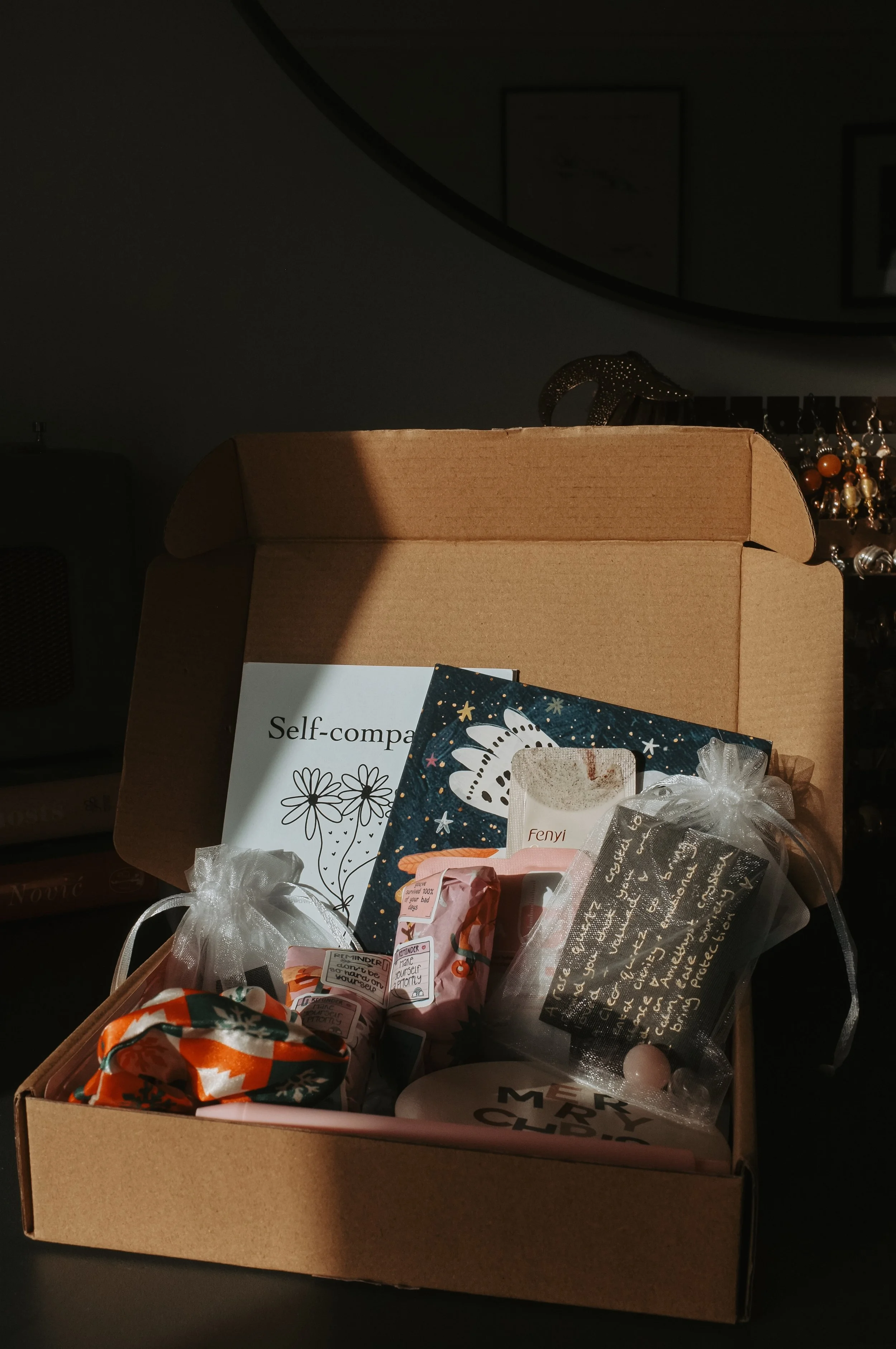 A cardboard box filled with wrapped gifts and cards, positioned on a dark surface with a mirror and jewelry in the background, illuminated by a beam of sunlight.
