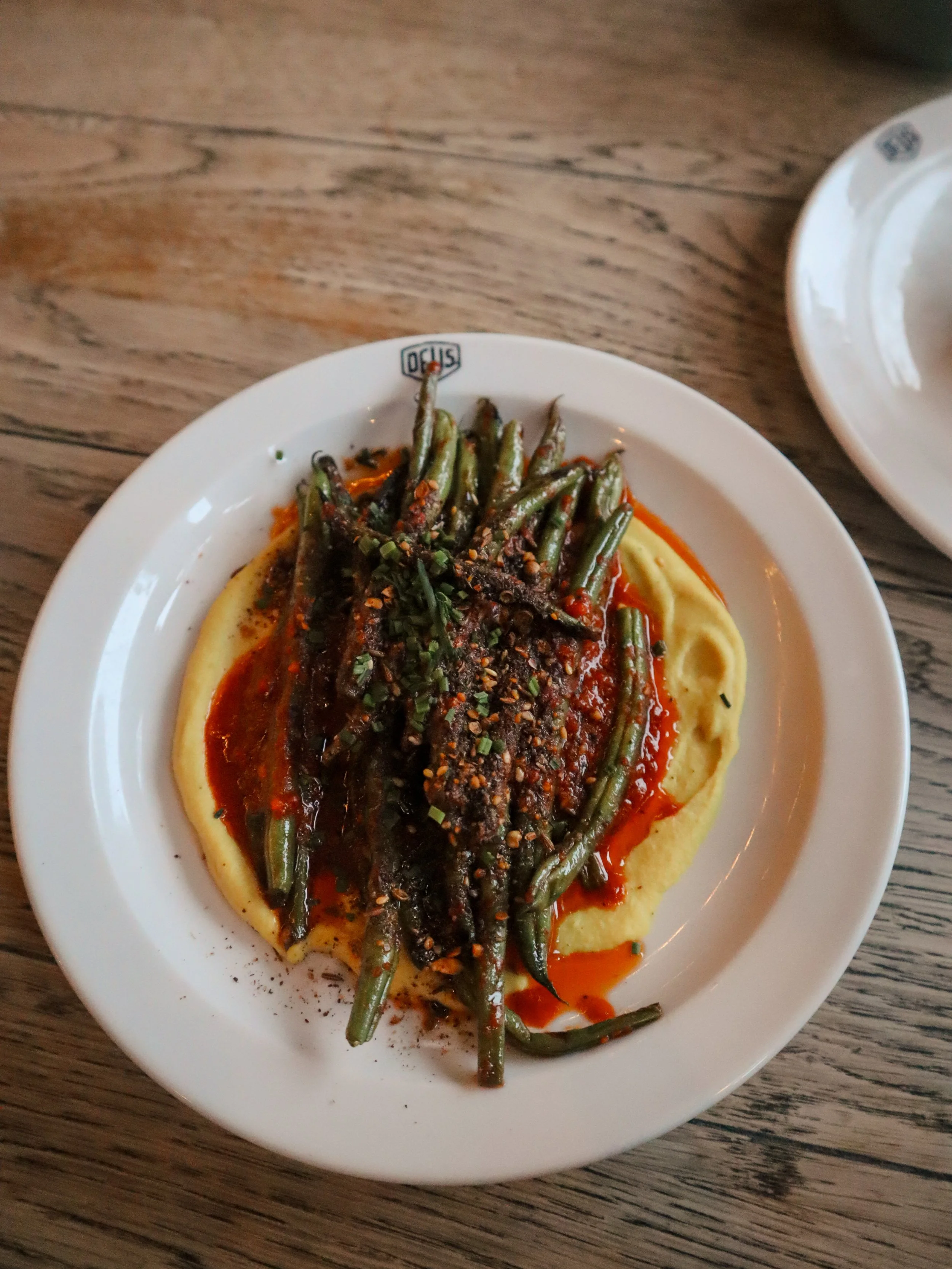 A white plate with steak topped with green beans and red sauce, served on mashed potatoes, placed on a wooden table.