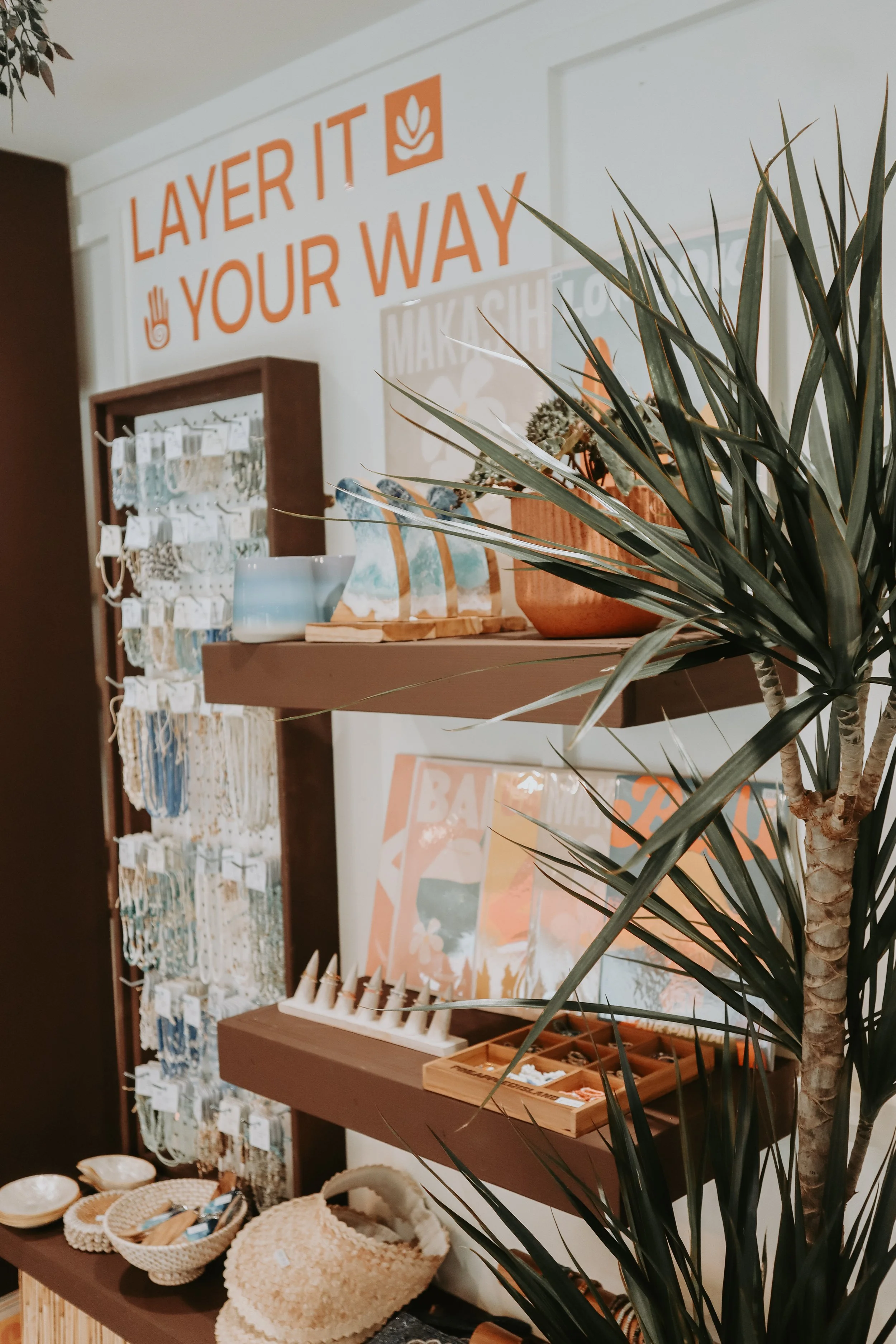 Display of various home decor items, jewelry, and accessories on shelves and tables in a retail store setting with a large sign that reads 'Layer it Your Way' in orange letters on a white background.