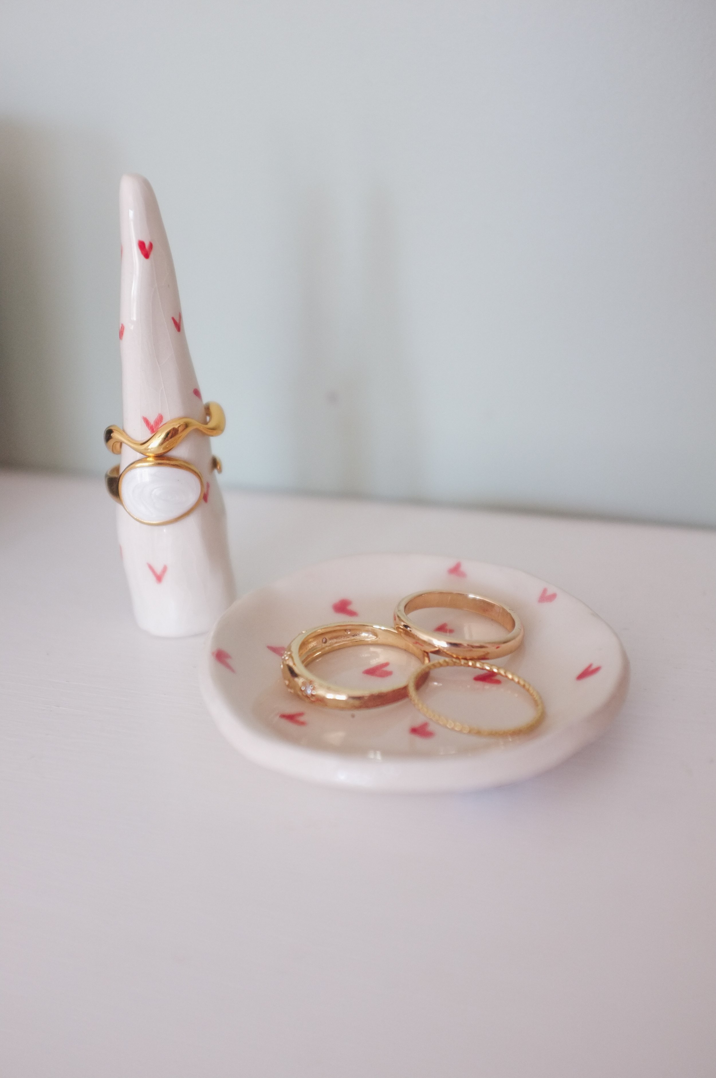 Close-up of a small white ceramic dish with pink hearts, containing three gold rings, in front of a ceramic unicorn figurine decorated with small pink hearts and wearing a gold ring around its neck, against a plain white background.