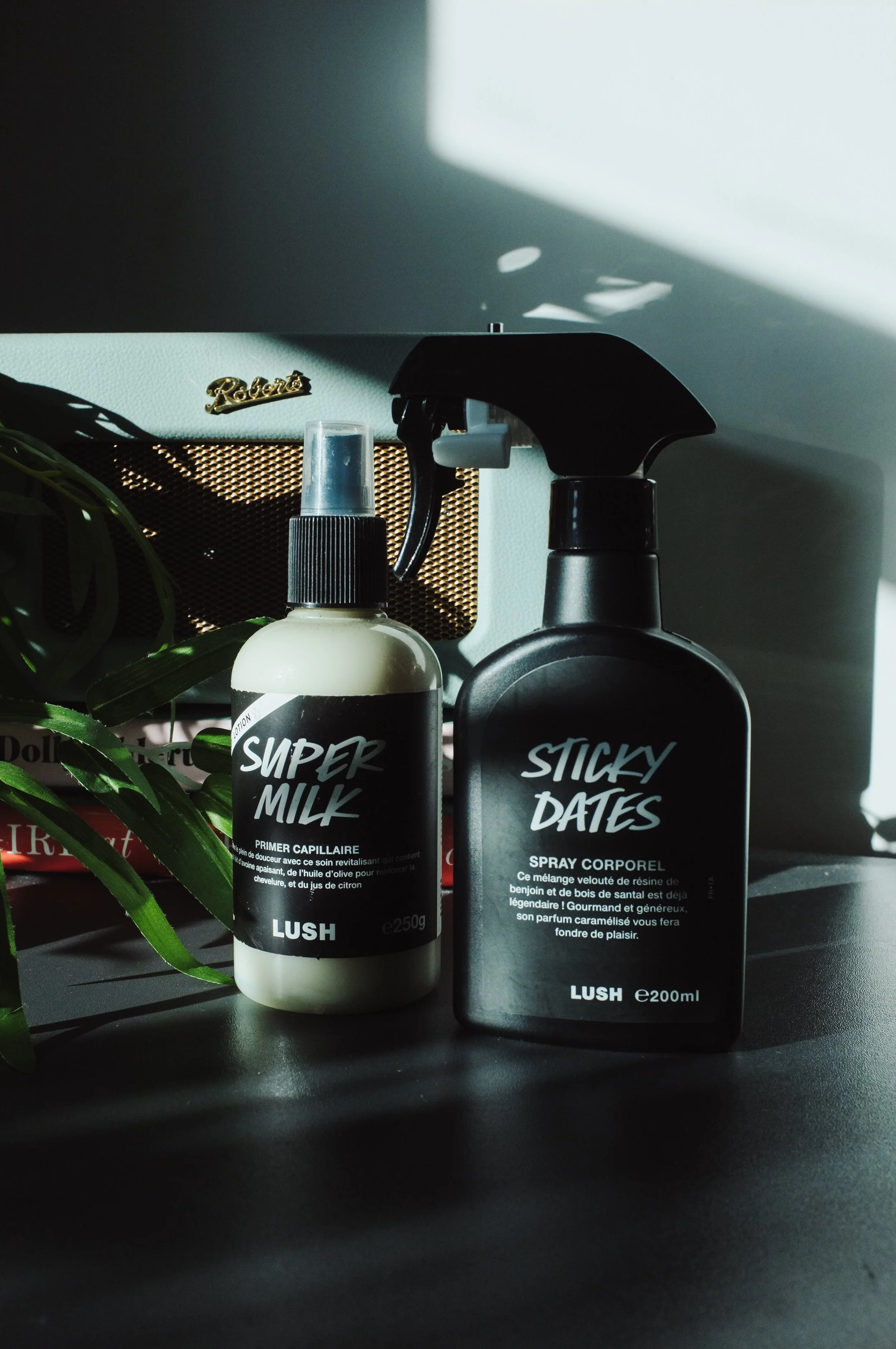 Two black and white bottles of Lush brand hair care products on a dark surface, with green leaves partially visible on the left and a laptop in the background. One bottle has a spray top and the other has a spray nozzle.