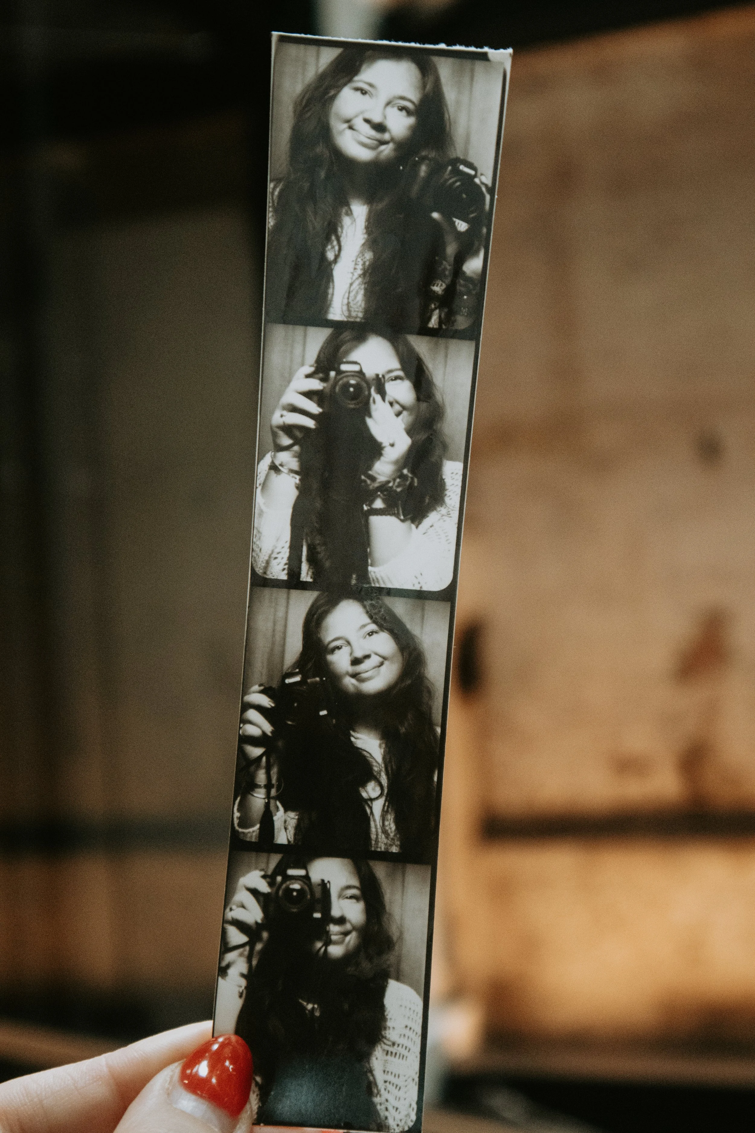 A strip of black-and-white photo booth pictures of a woman with long, wavy hair, smiling and holding a camera. The woman appears in all frames, sometimes covering her face with the camera and other times smiling at the camera.