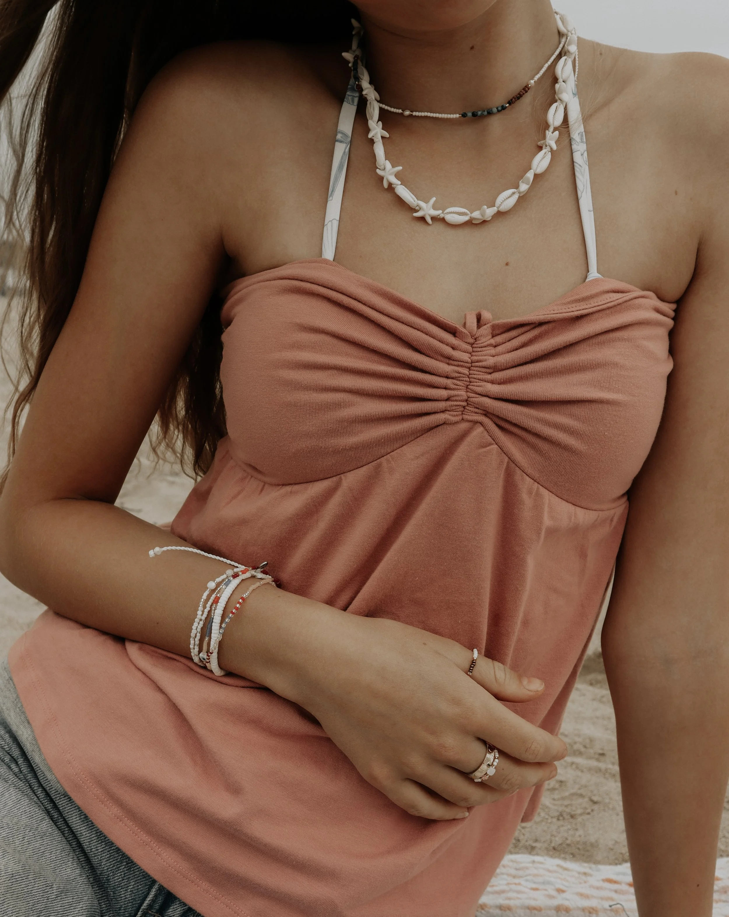 A woman wearing a peach-colored strapless top, layered necklaces with shells and beads, and bracelets, standing outdoors on a sandy area.
