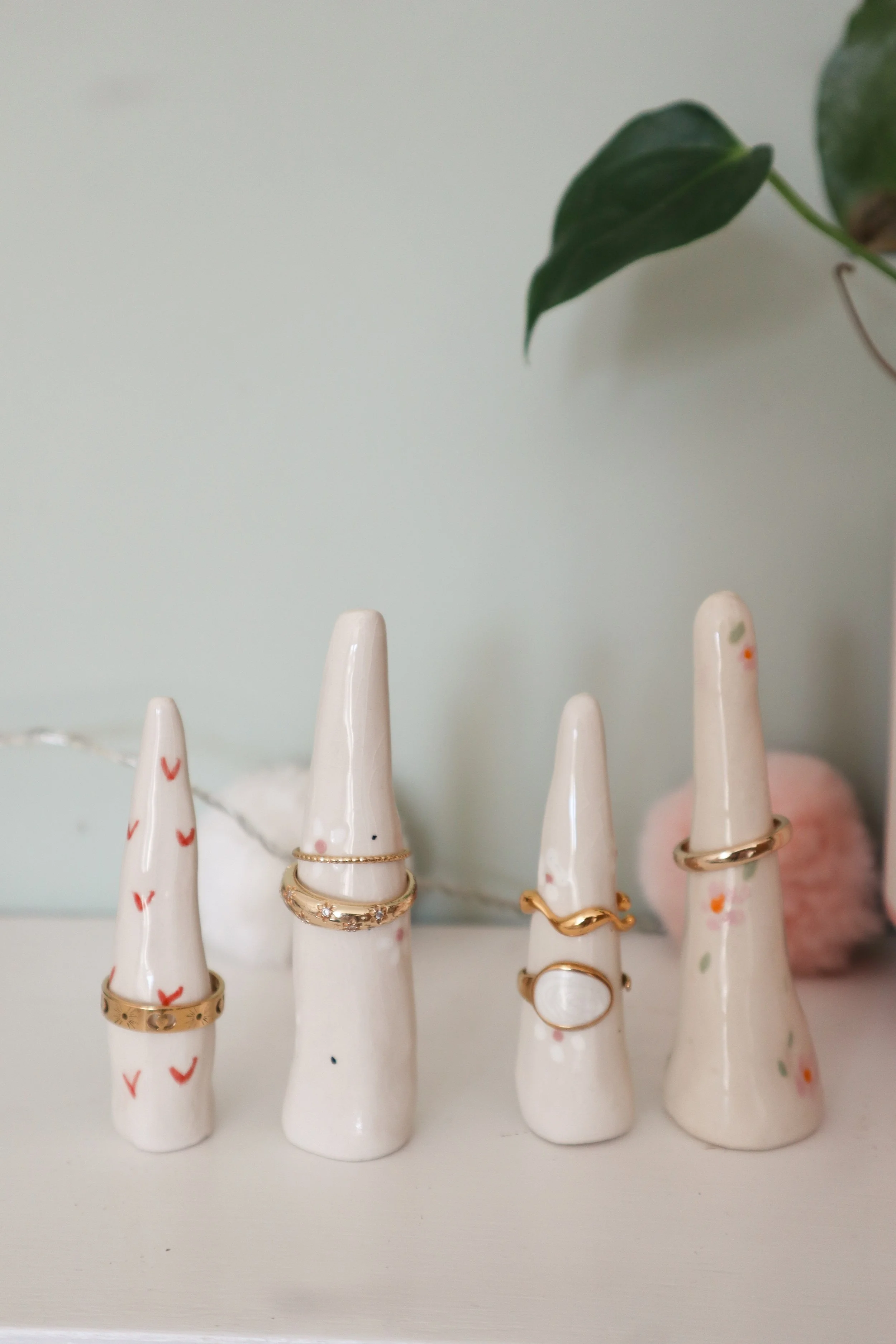 Multiple white ceramic ring holders in the shape of stylized fingers, adorned with various gold and silver rings, with a background of a light-colored wall and a green leaf.