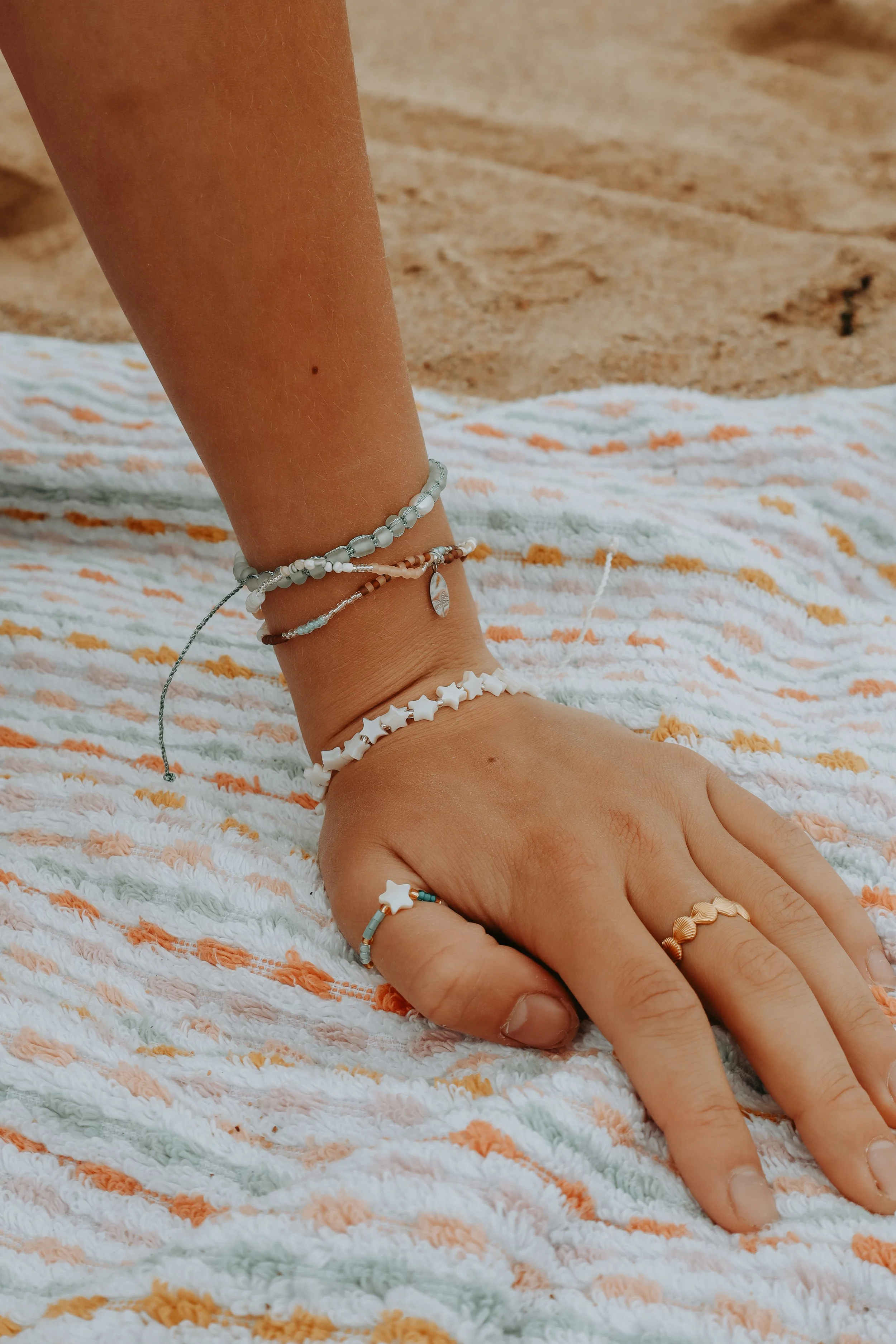 A person's arm resting on a multicolored towel on a sandy beach, adorned with various beaded and shell jewelry.