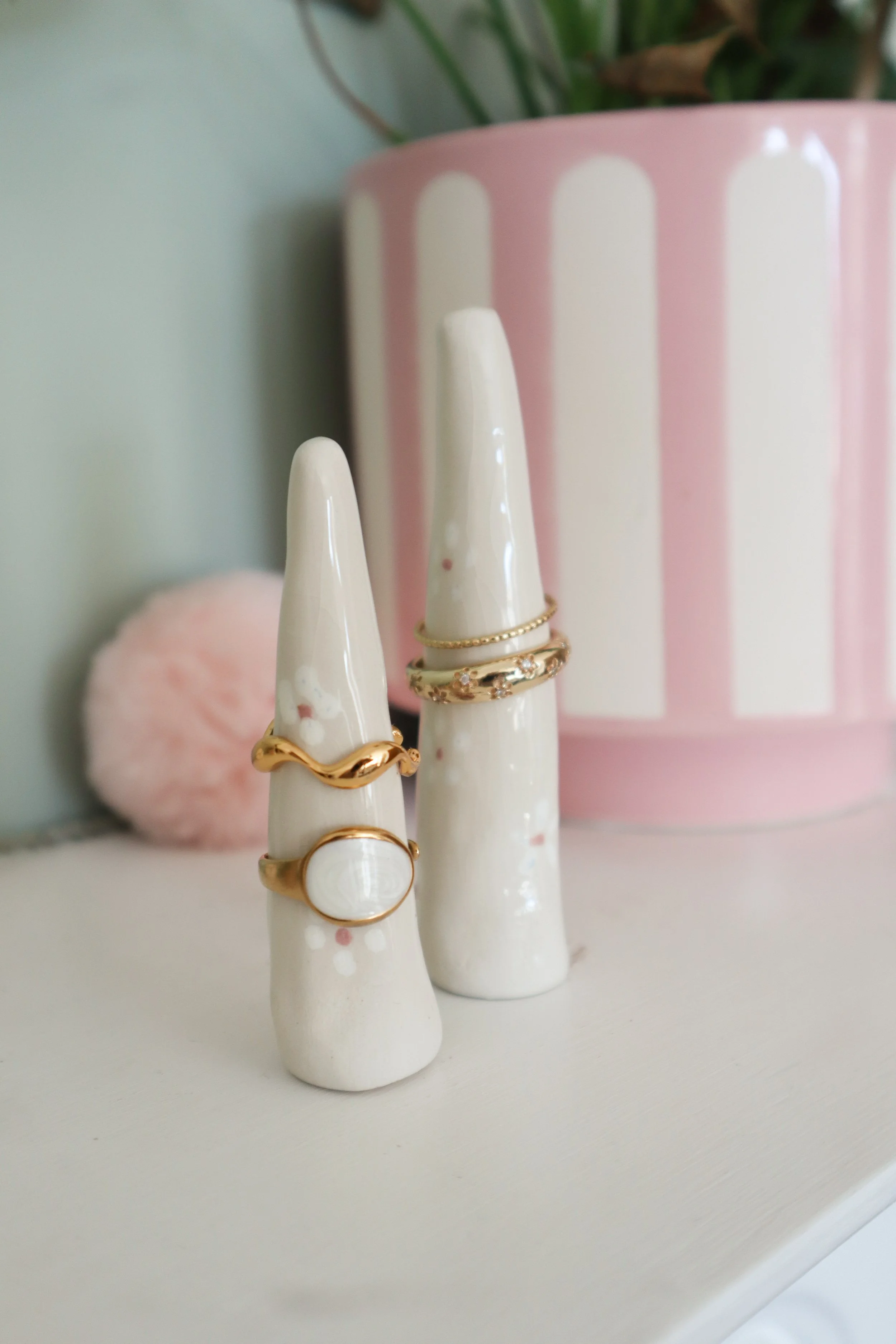 Three ceramic bunny-shaped ring holders displaying gold rings with stones, against a pink and white striped ceramic vase on a white surface.