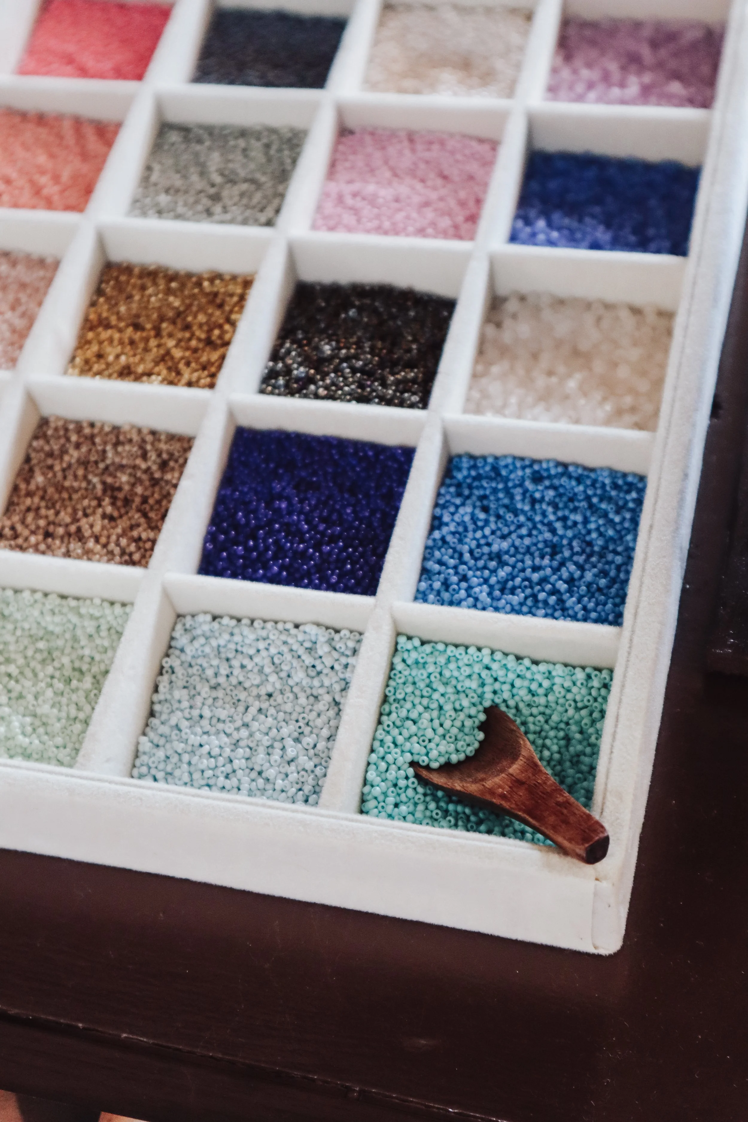 A white tray with multiple compartments containing colorful small beads, and a wooden spoon resting in one compartment. Pineapple Island Jewellery Photography