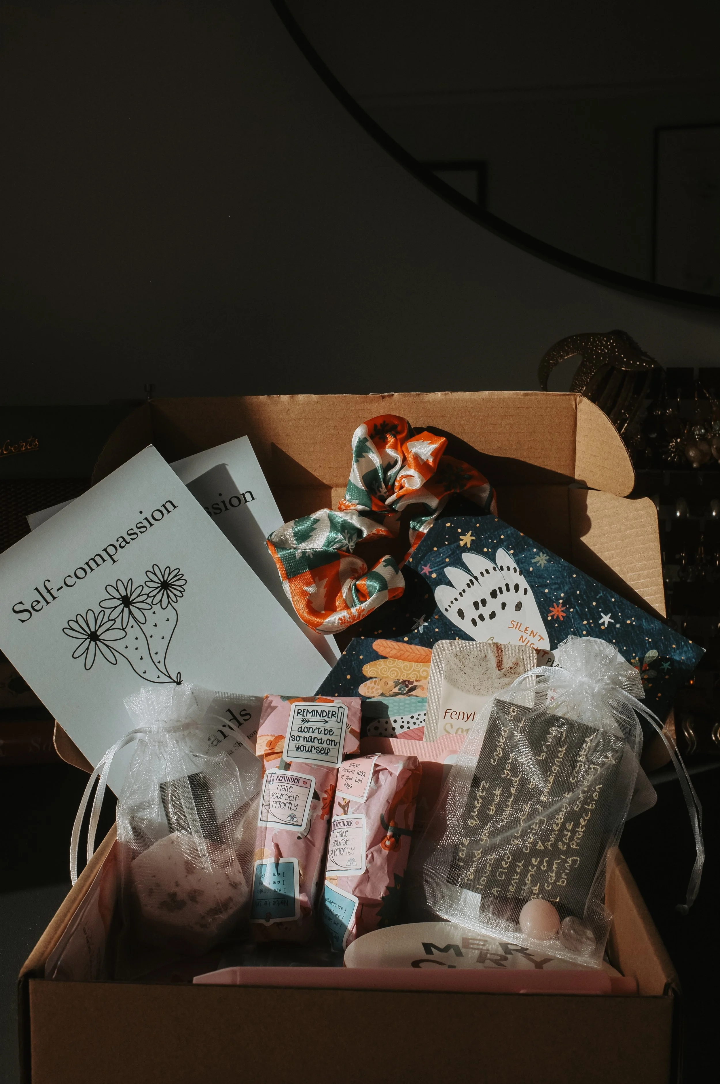 Open cardboard box filled with various small gifts, cards, and decorative items, including a card titled 'Self-compassion,' wrapped packages, and a silk scrunchie.