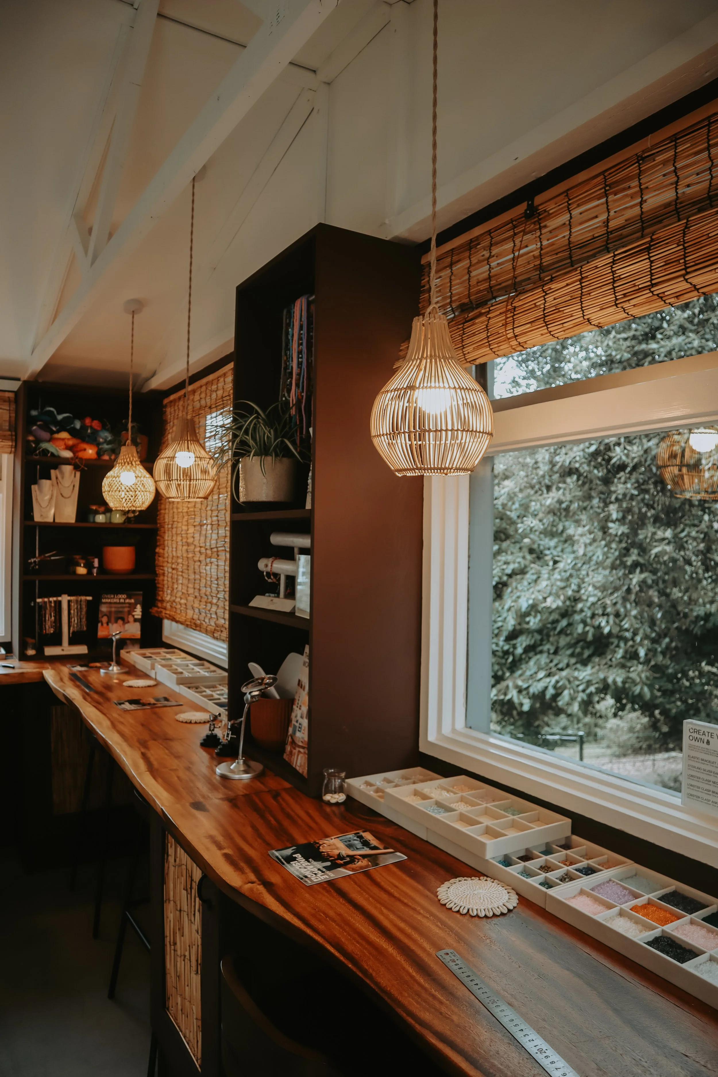 Interior of a craft or jewelry store with a wooden counter, display trays with various beads or small materials, hanging woven light fixtures, a window showing trees outside, and shelves with books and decorations.