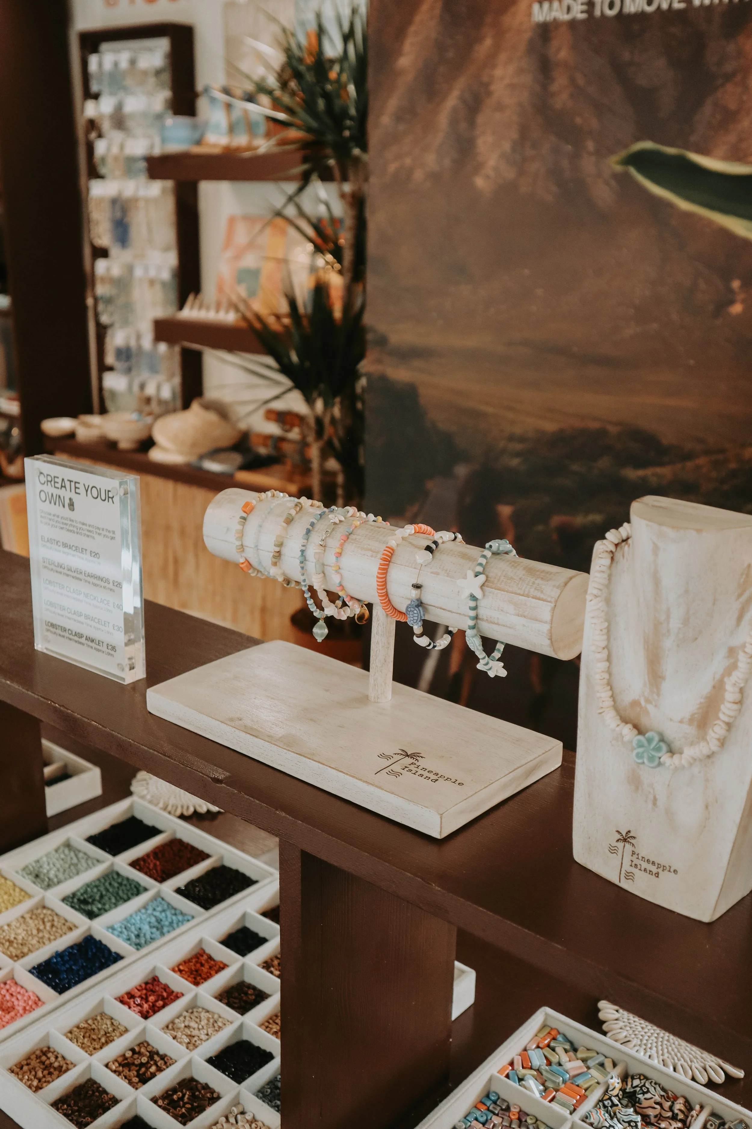Display of colorful beaded jewelry on a wooden stand, with a sign reading "Create Your Own" and a background of a desert landscape with mountains and cacti. Pineapple Island Jewellery Photography