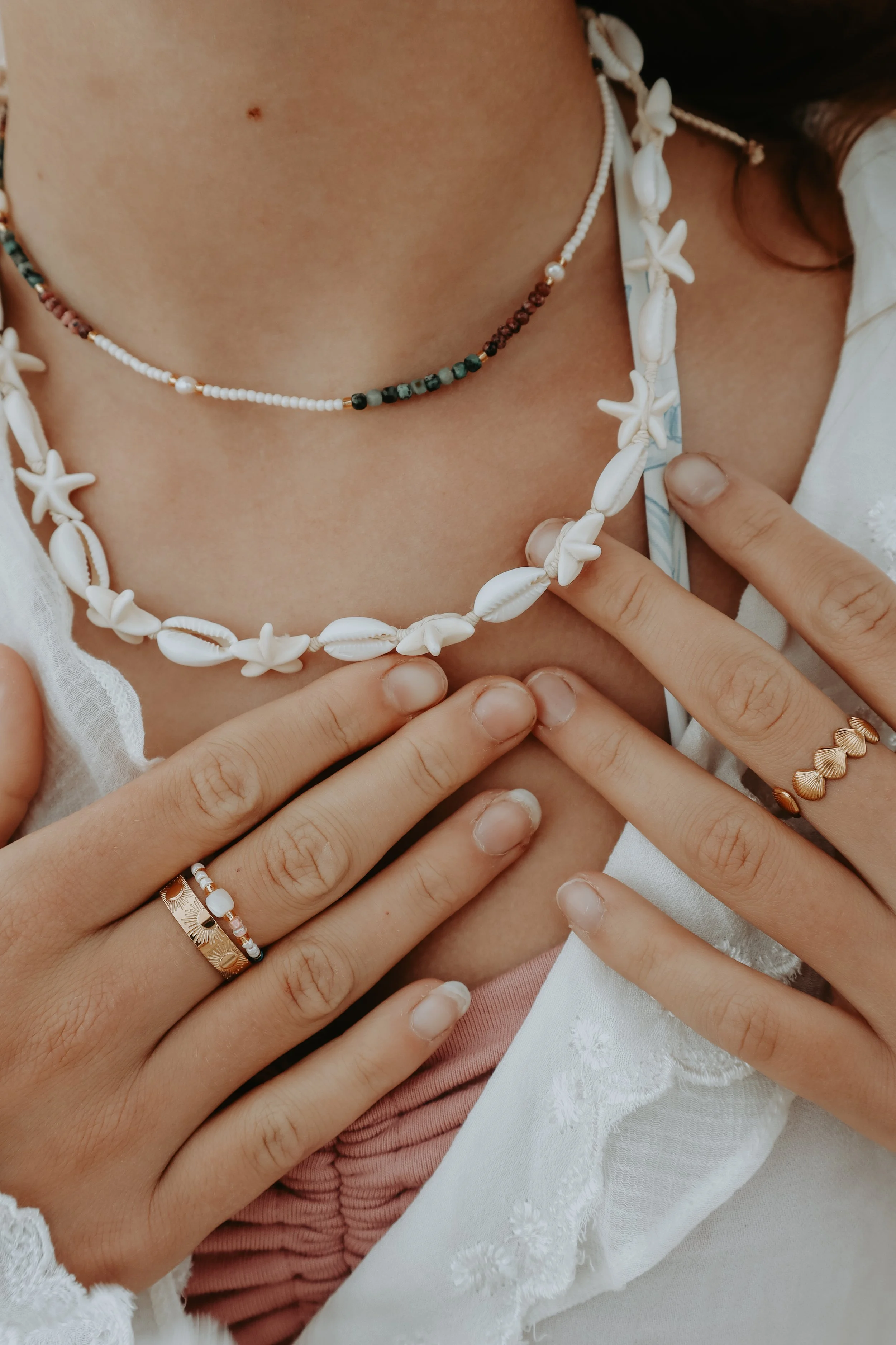Close-up of a person's neck and hands, featuring layered necklaces made of white beads and shells, and fingers with rings, over white fabric with floral embroidery.