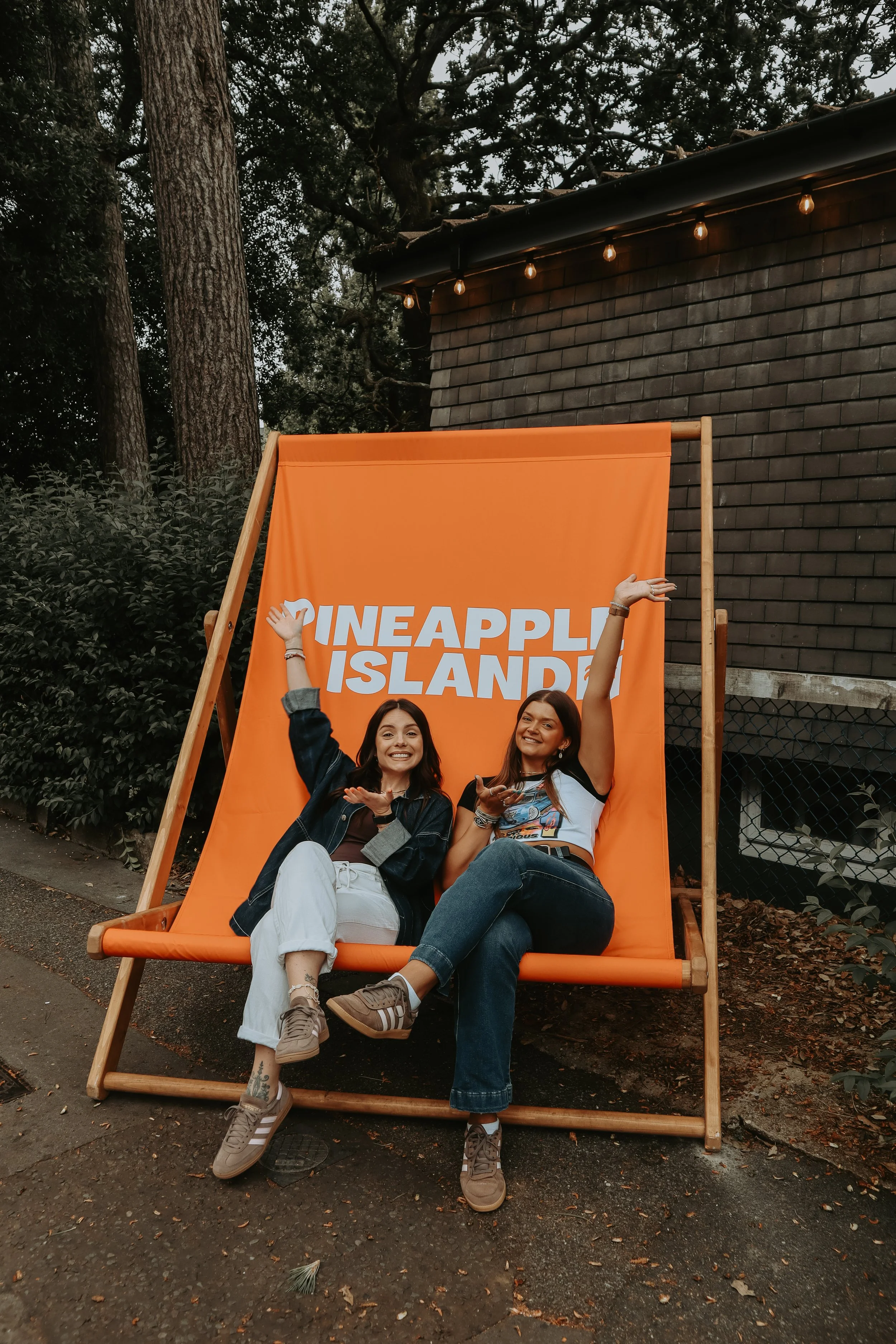 Two women sitting on an orange outdoor lounge chair in front of an orange sign with white text that reads 'Pineapple Island.' One woman has dark hair, wears a denim jacket, white pants, and sneakers, and is making a peace sign. The other woman has da