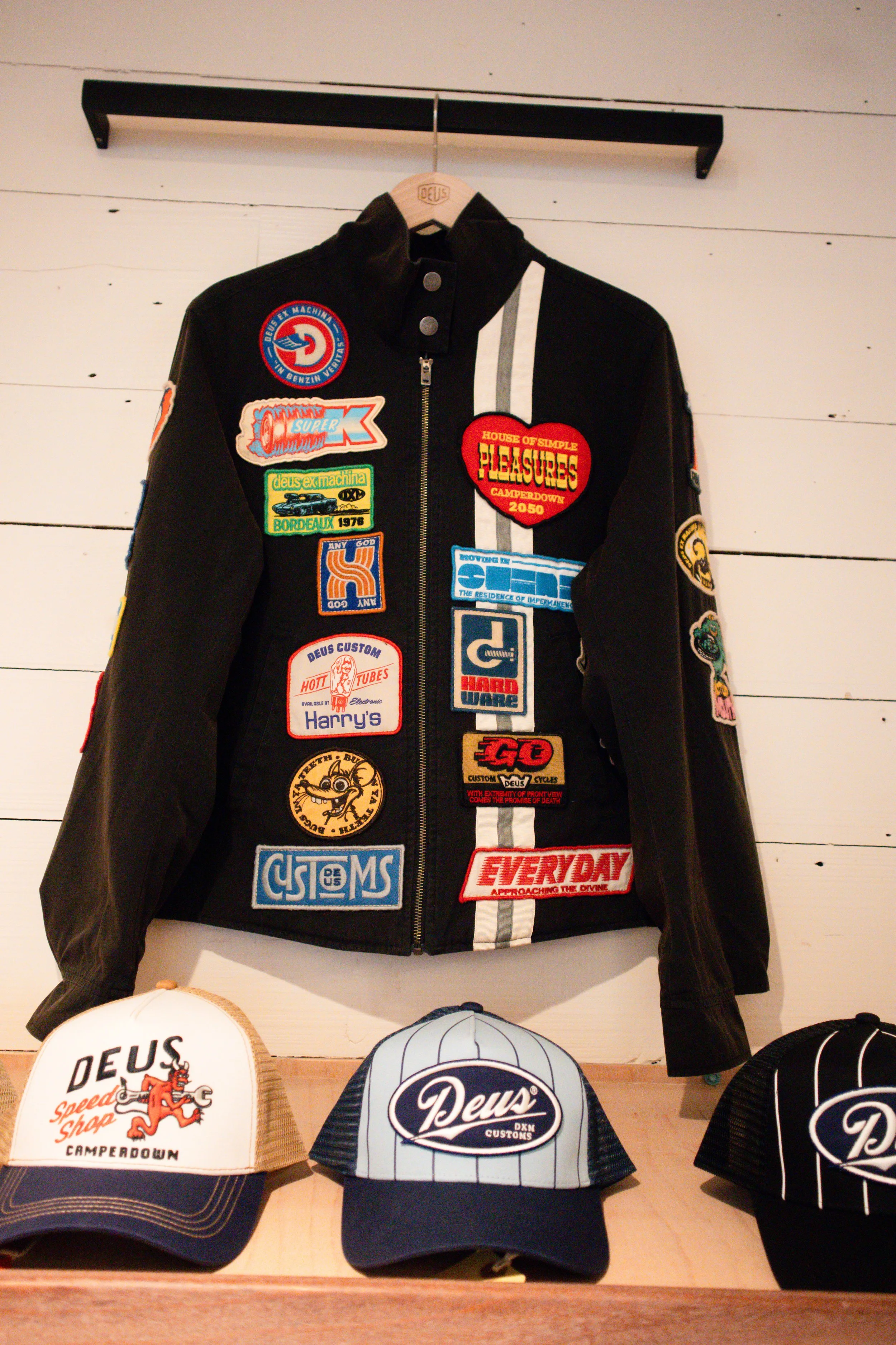 A black leather jacket with various colorful patches hanging on a hanger, displayed on a wall, next to a display of baseball caps with different logos and designs.