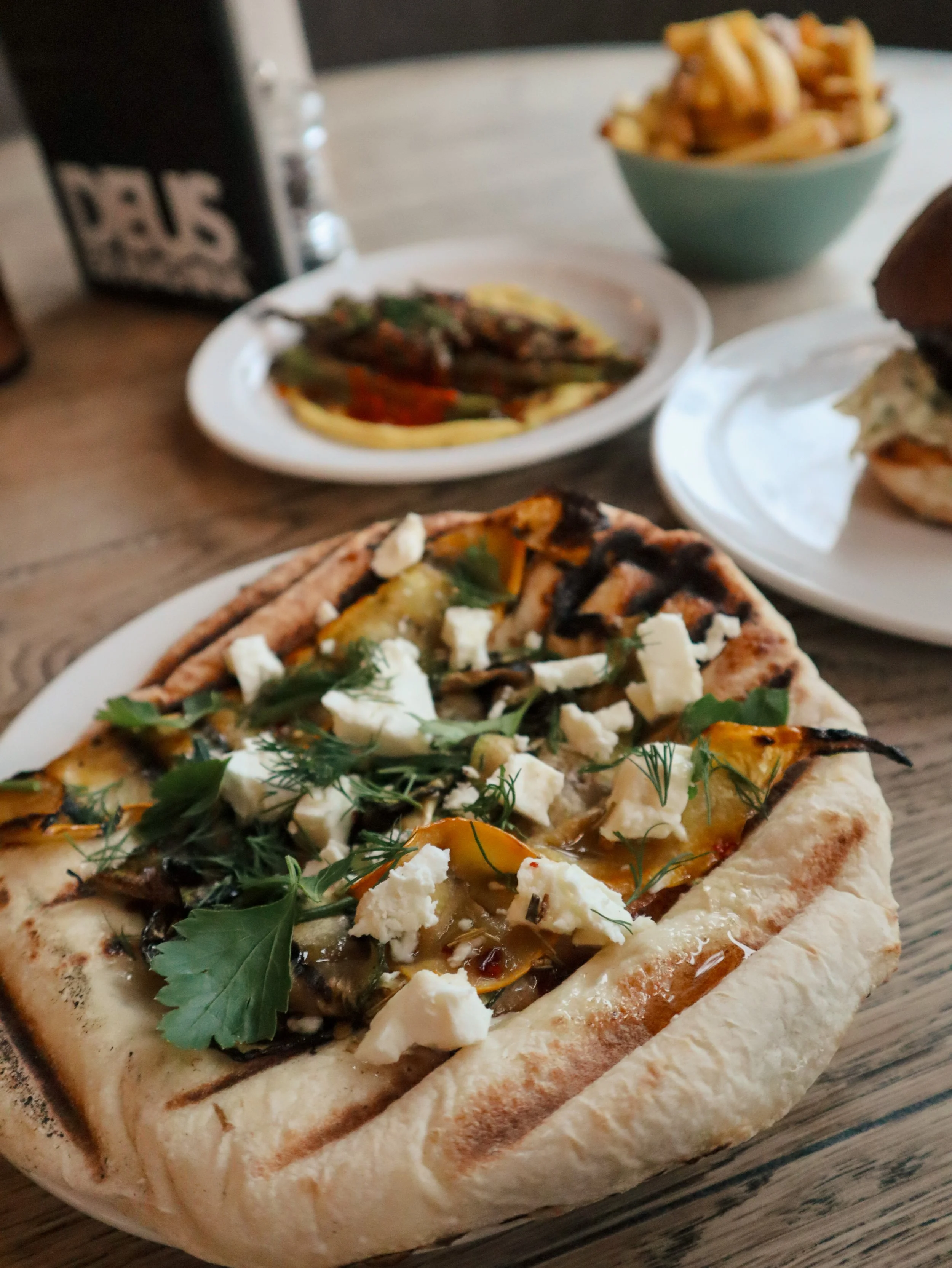 Close-up of a grilled vegetable flatbread pizza topped with herbs, cheese, and vegetables, on a wooden surface.