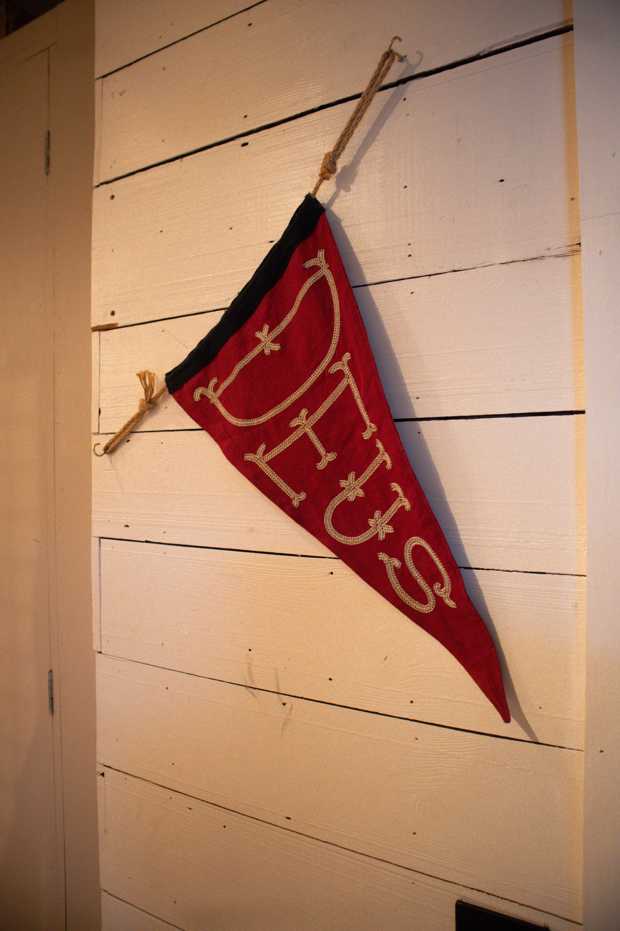 Red fabric banner with embroidered gold lettering hanging on a white wooden wall with horizontal planks. The banner is attached to the wall with a thick rope and metal hooks.