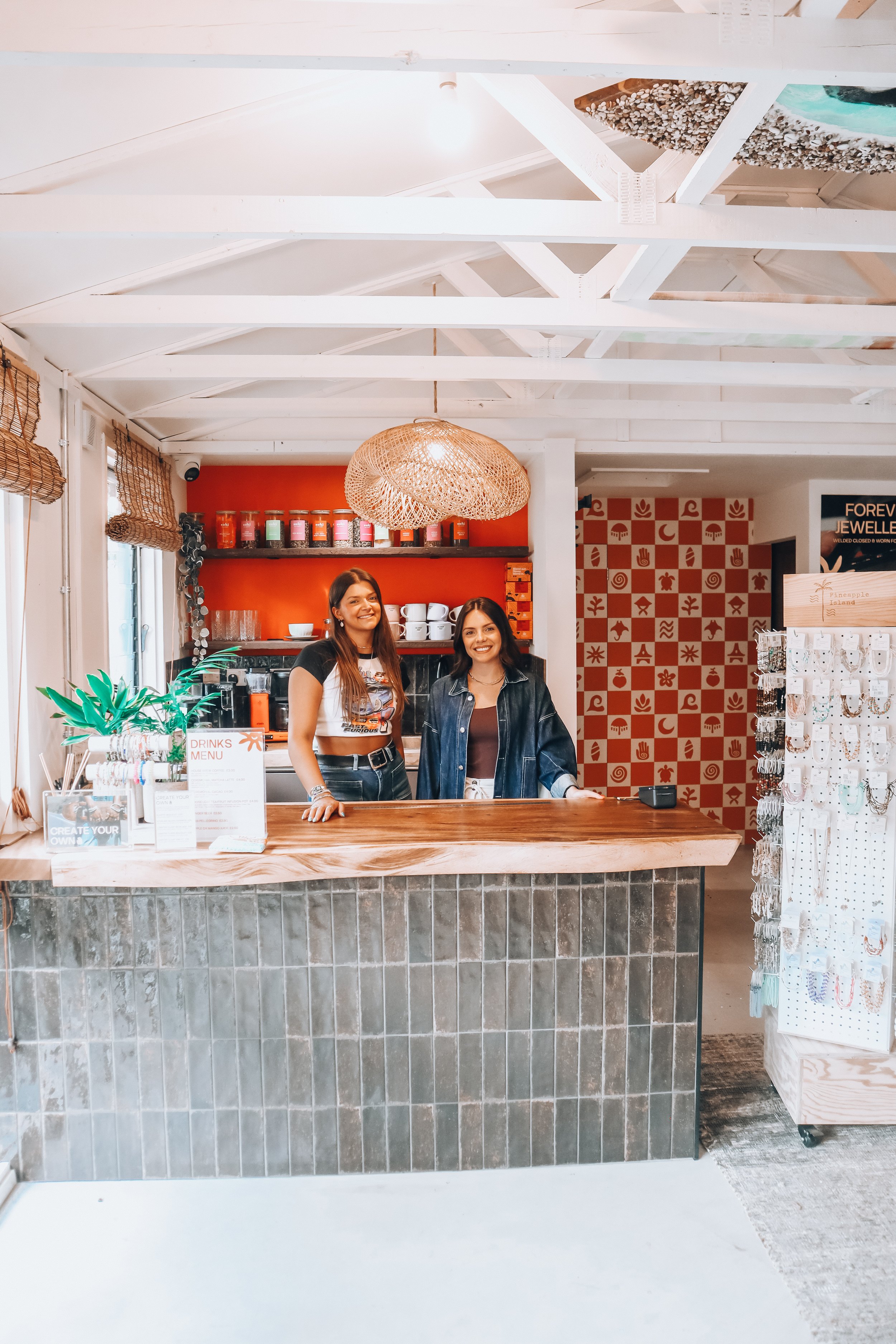 Two women standing behind a colorful wooden counter in a modern, brightly decorated shop or cafe, with one woman wearing a graphic T-shirt and the other in a denim jacket, smiling at the camera. The background features orange and red walls, a shelf w