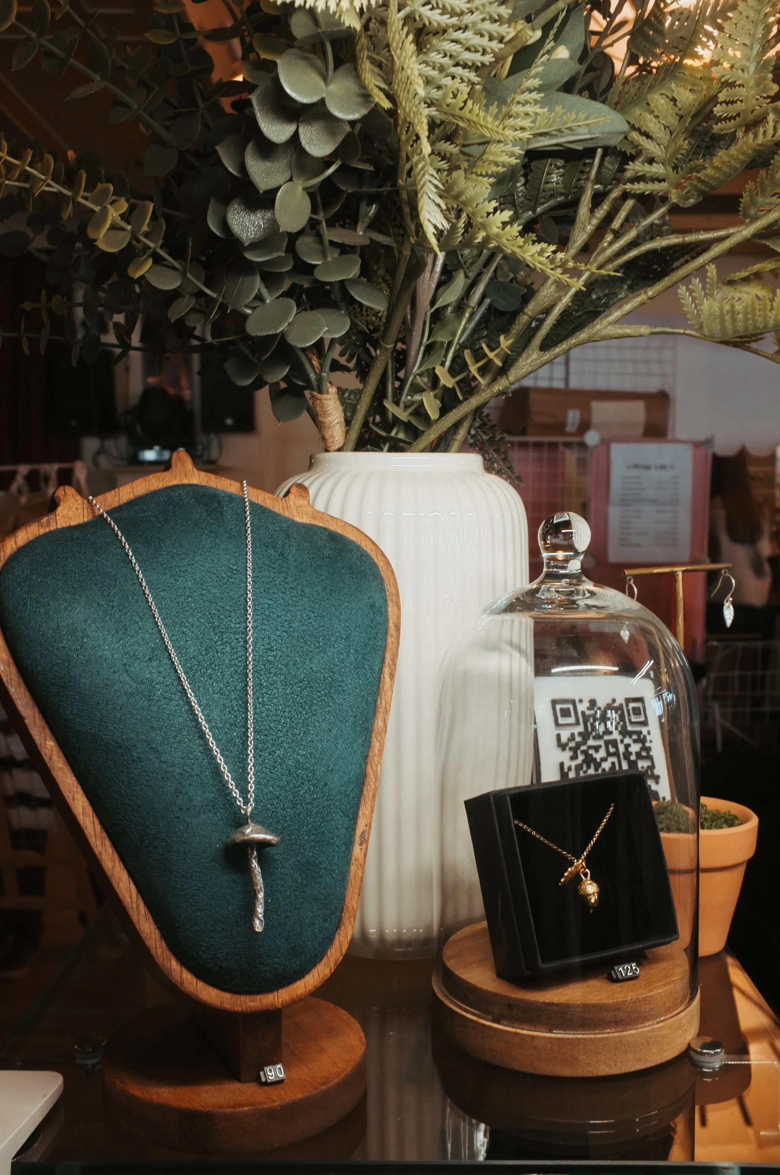 Jewelry display with necklaces inside a glass dome and on a green display bust, against a large arrangement of green foliage in a white vase.